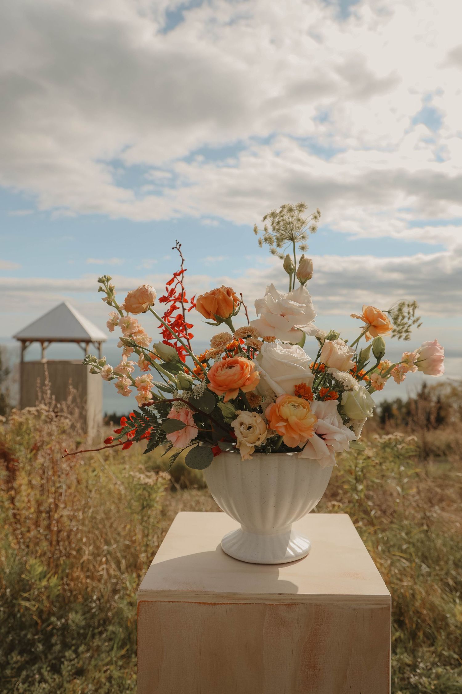 Elegant white vase with peach and orange floral arrangement on pedestal against cloudy sky outdoors.