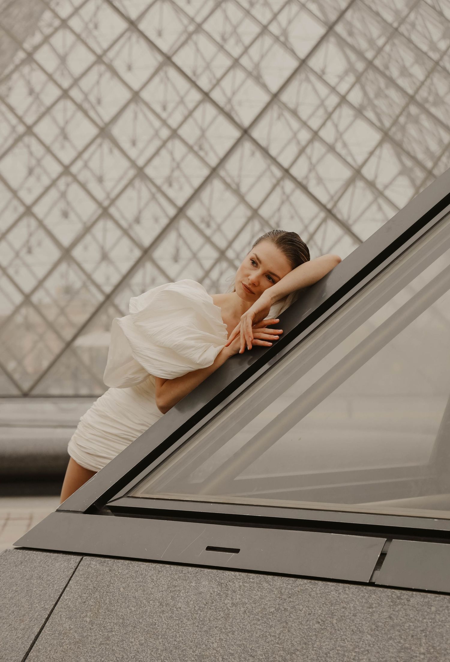 Woman in white puff-sleeve mini dress leans gracefully against the Louvre glass pyramid in Paris.