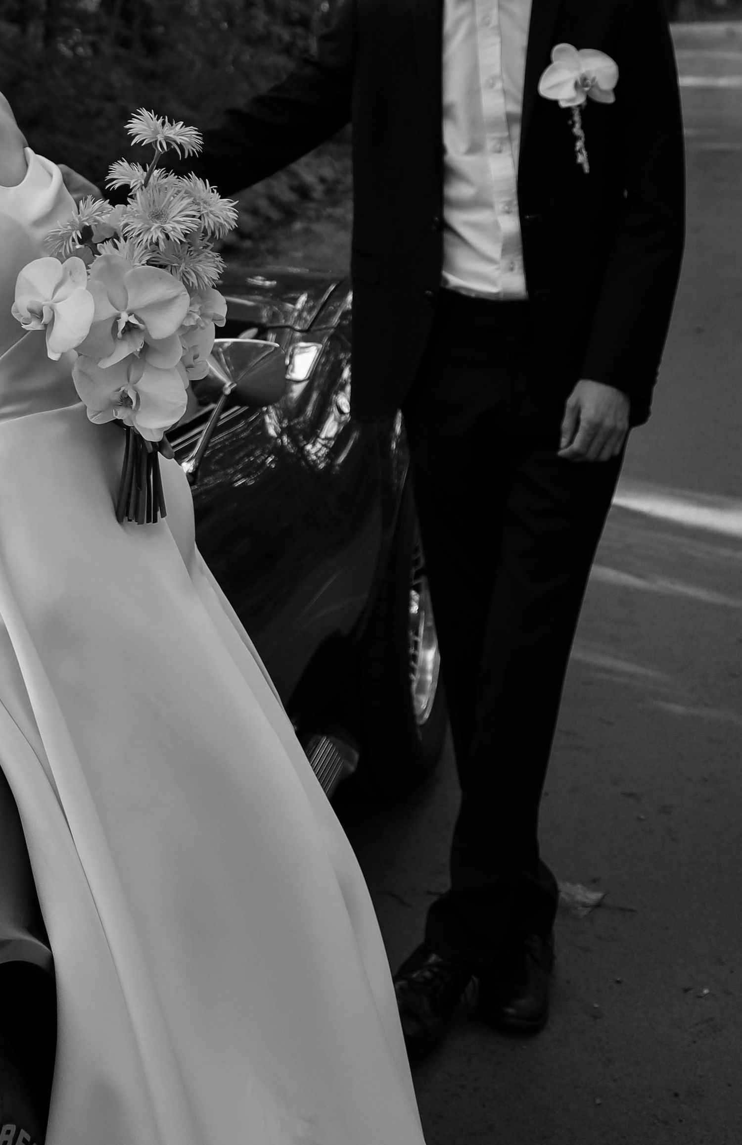 Black and white photo of bride holding bouquet and groom in tuxedo standing beside classic car on wedding day.