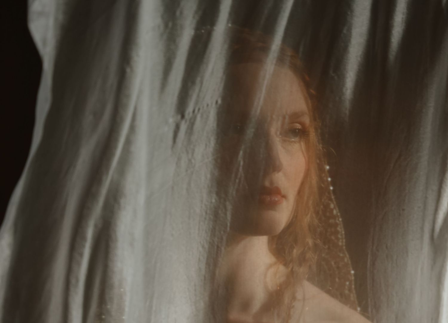 Ethereal bride with red hair seen through sheer white fabric curtain in moody lighting.