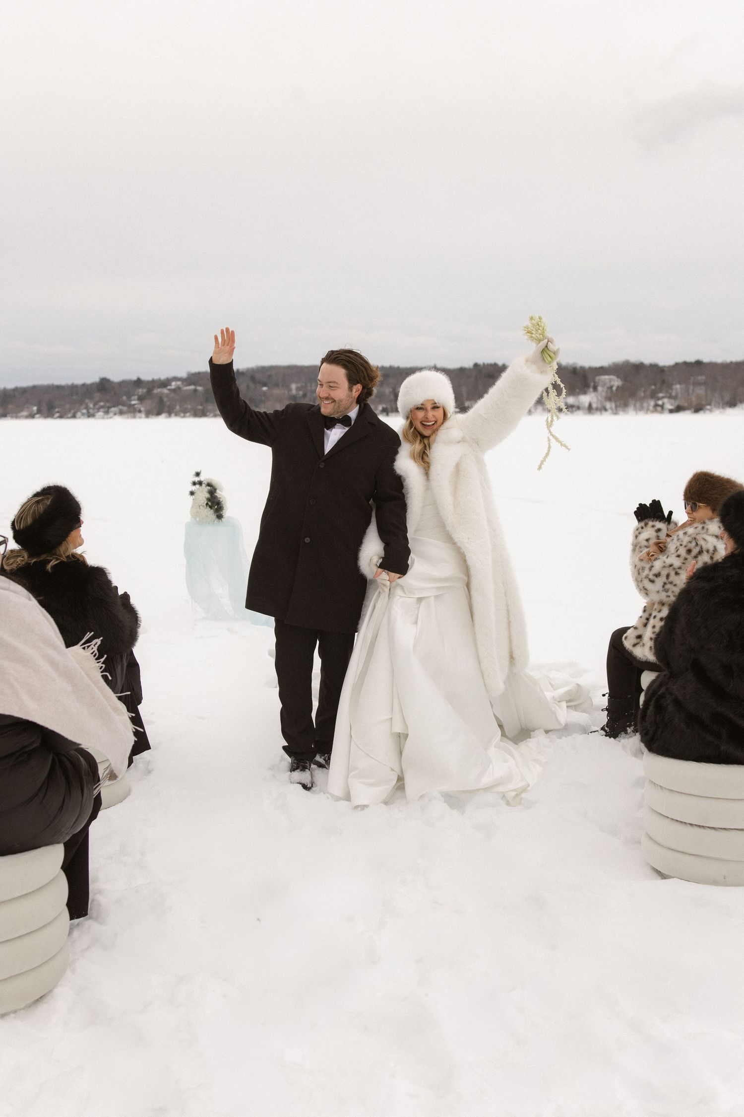 Happy couple raising hands after outdoor winter wedding ceremony on snowy lake surrounded by guests.