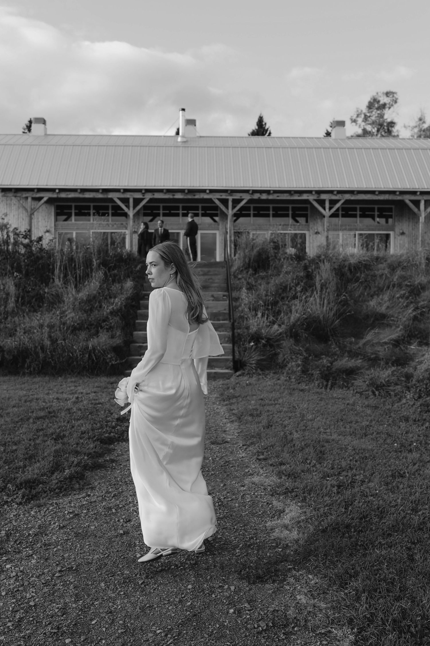 Bride in elegant white long-sleeve gown walking toward rustic wedding venue in black and white.