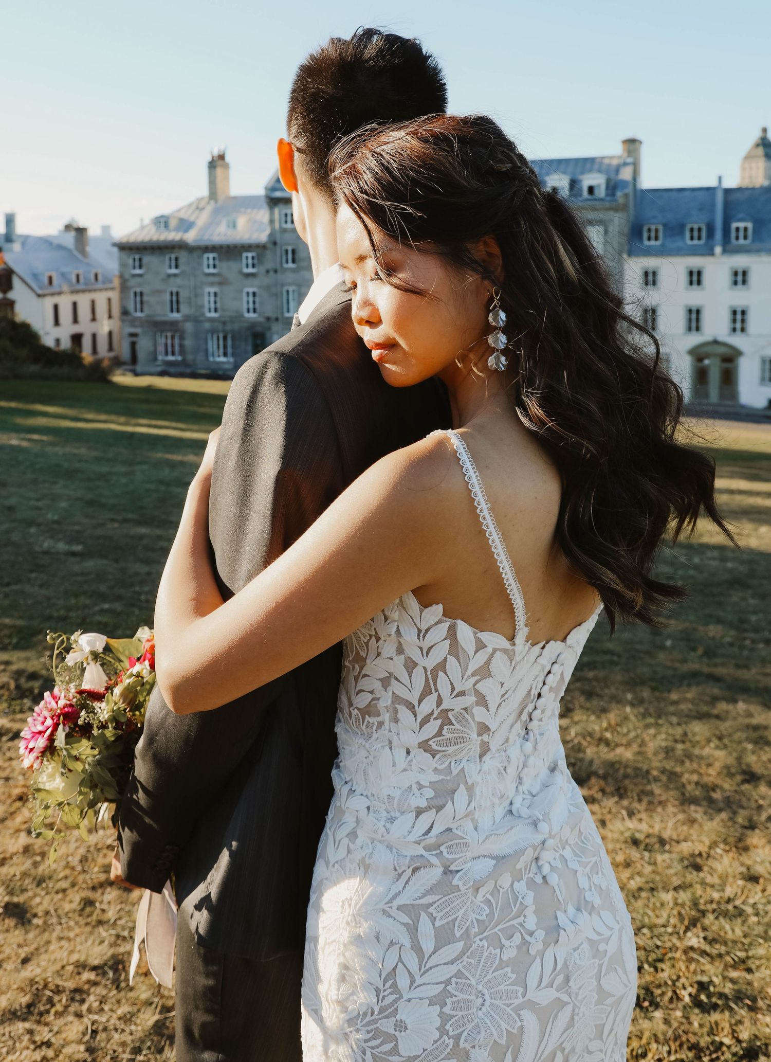 Bride in lace gown with floral bouquet leans on groom facing a historic stone manor estate.