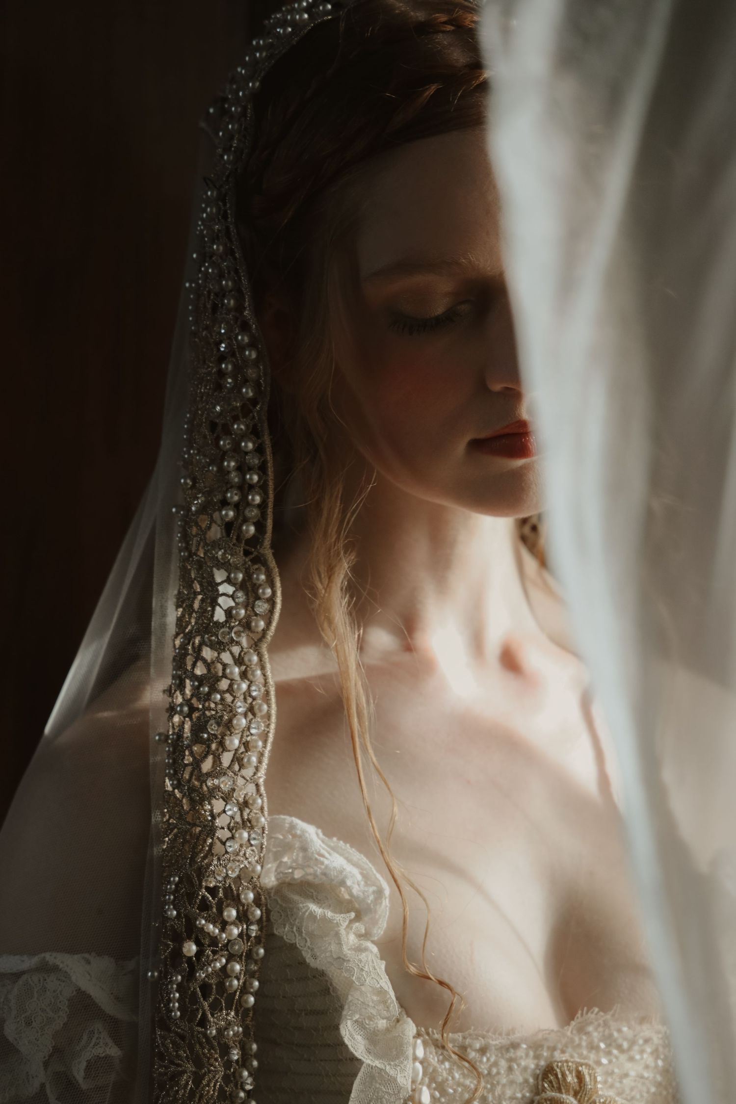 Bride in ornate beaded veil and lace wedding dress with eyes closed in soft dramatic lighting.