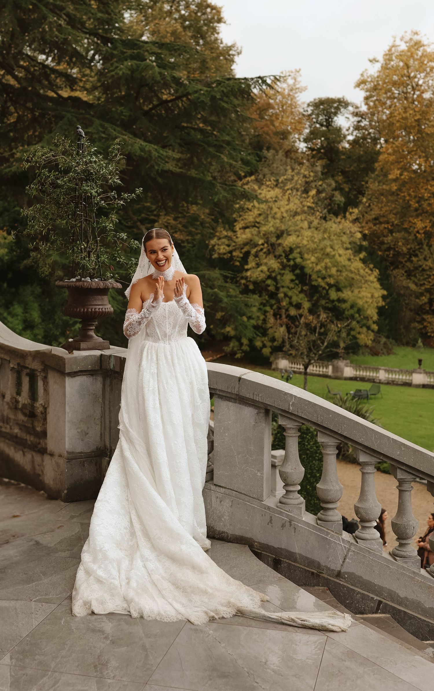 Stunning bride in a flowing lace wedding gown with veil posing on a stone balcony surrounded by autumn trees.