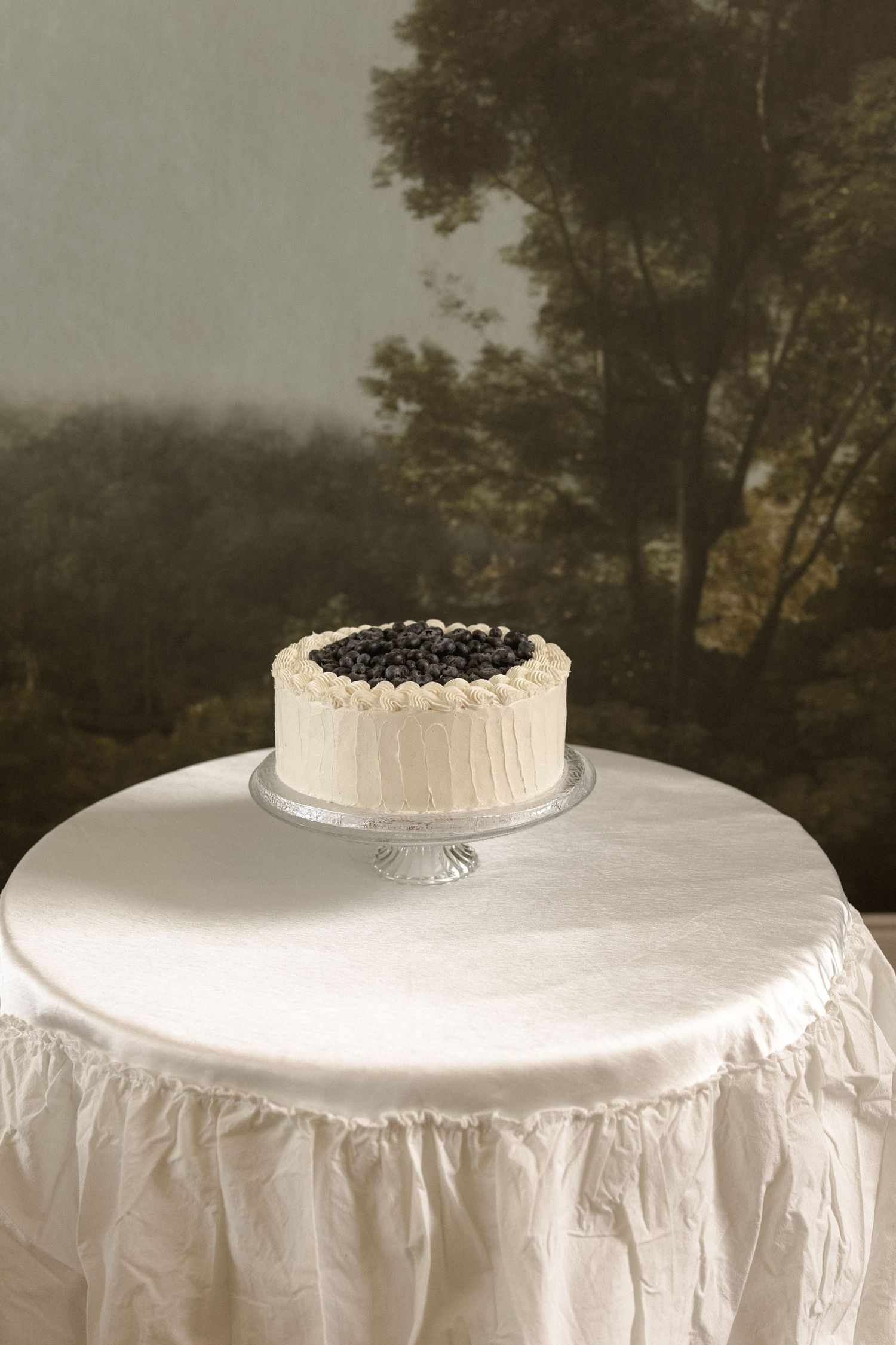 Simple white frosted wedding cake topped with fresh blueberries on glass stand with ruffled linen.