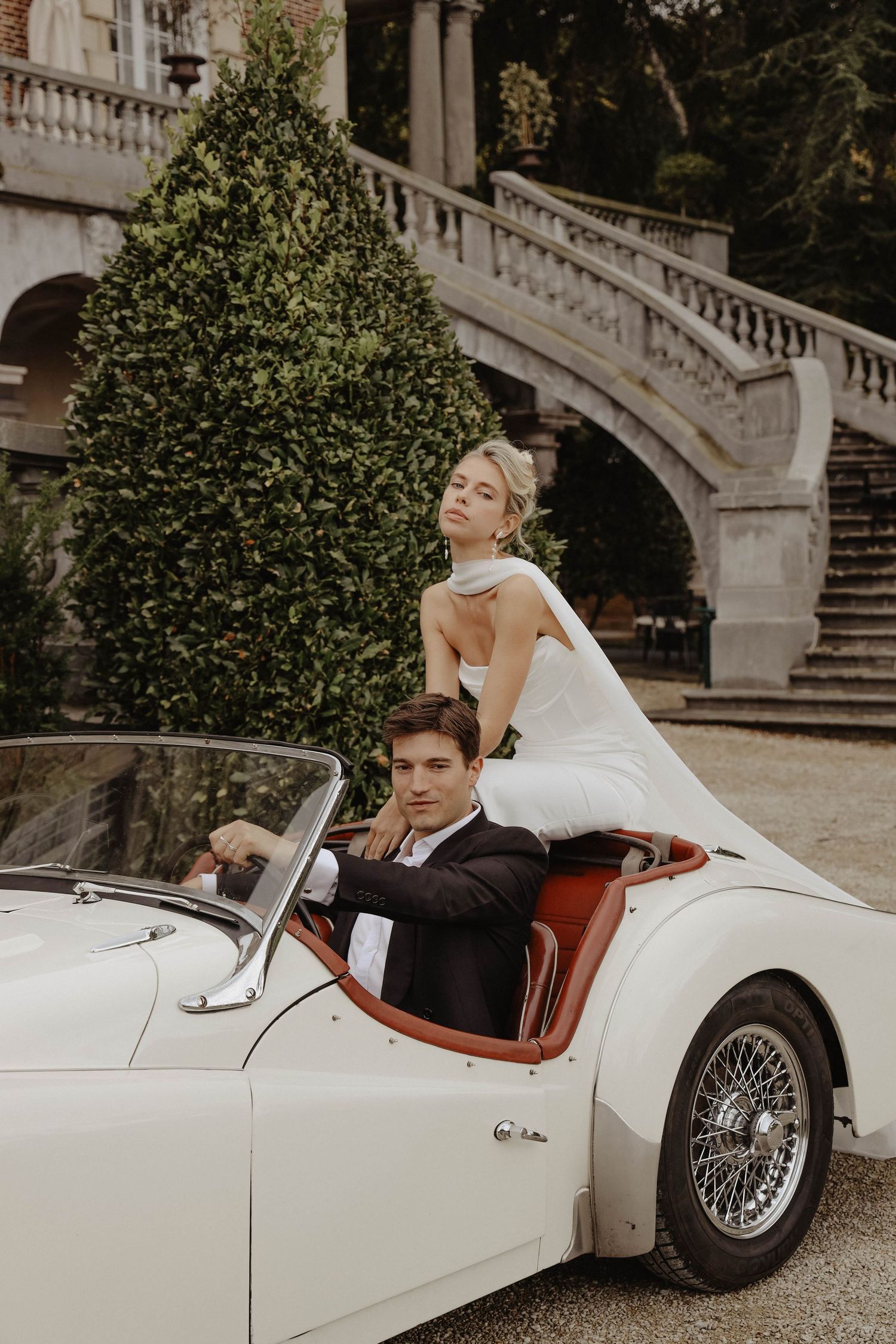 Bride stands behind groom seated in a vintage white Triumph convertible outside an elegant French chateau.