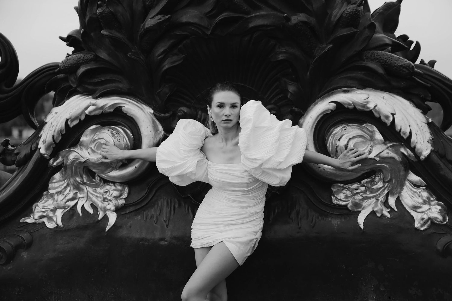 Black and white fashion photo of woman in white ruffled dress posed against ornate dark sculpture.