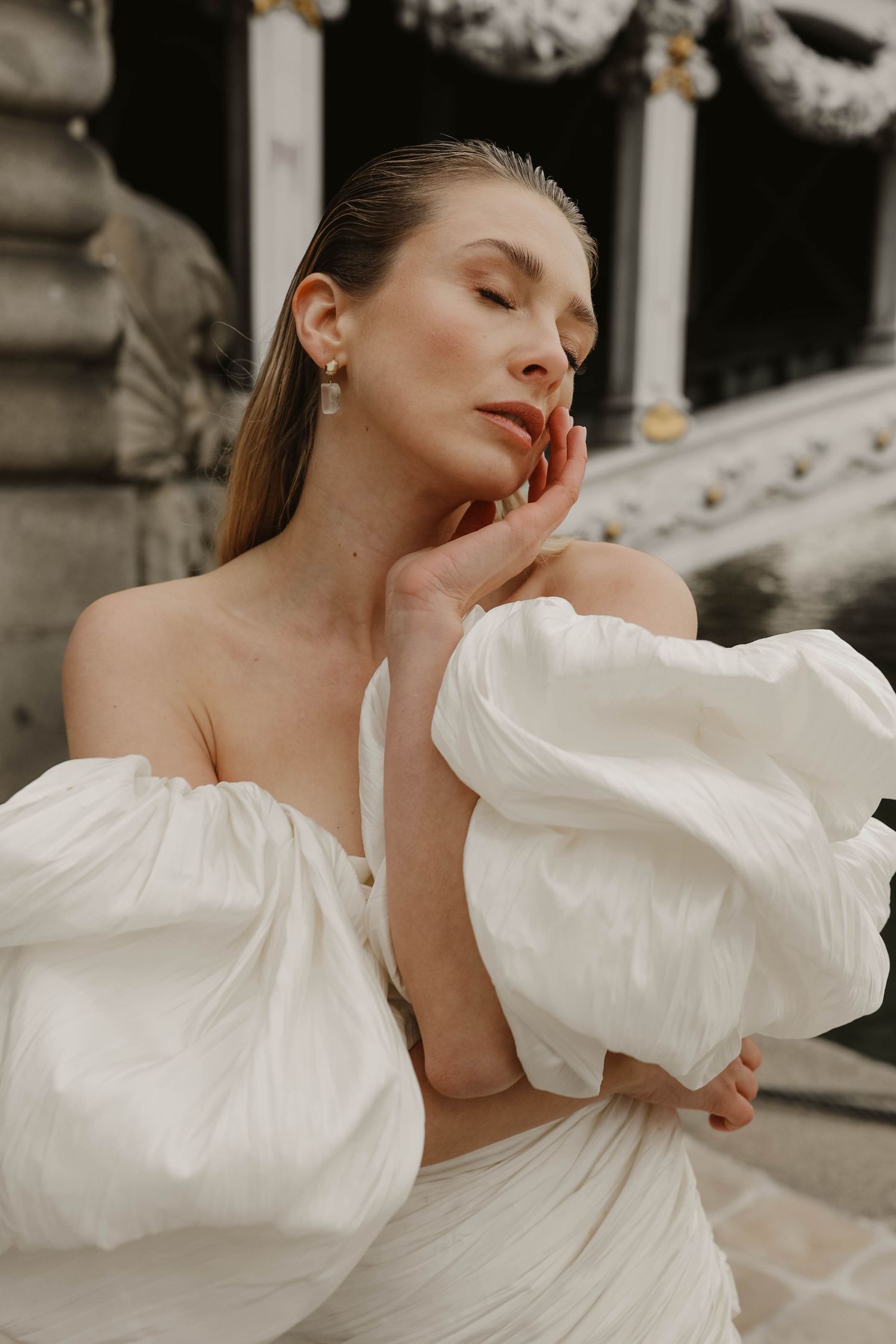 Woman in off-shoulder white puff-sleeve bridal gown posing with eyes closed near stone columns.