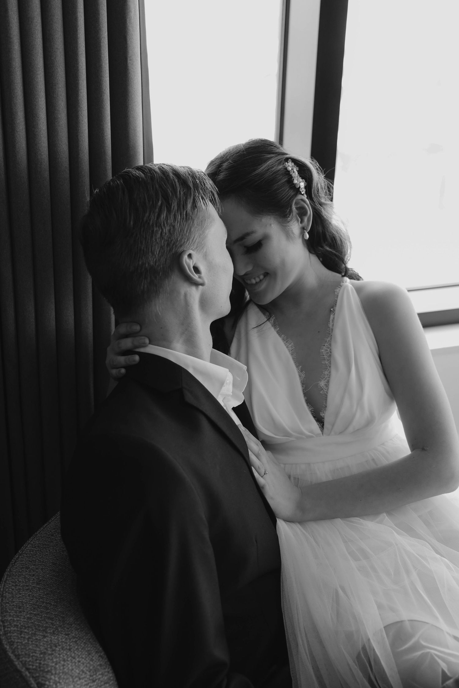 Black and white romantic wedding photo of smiling bride embracing groom by window.