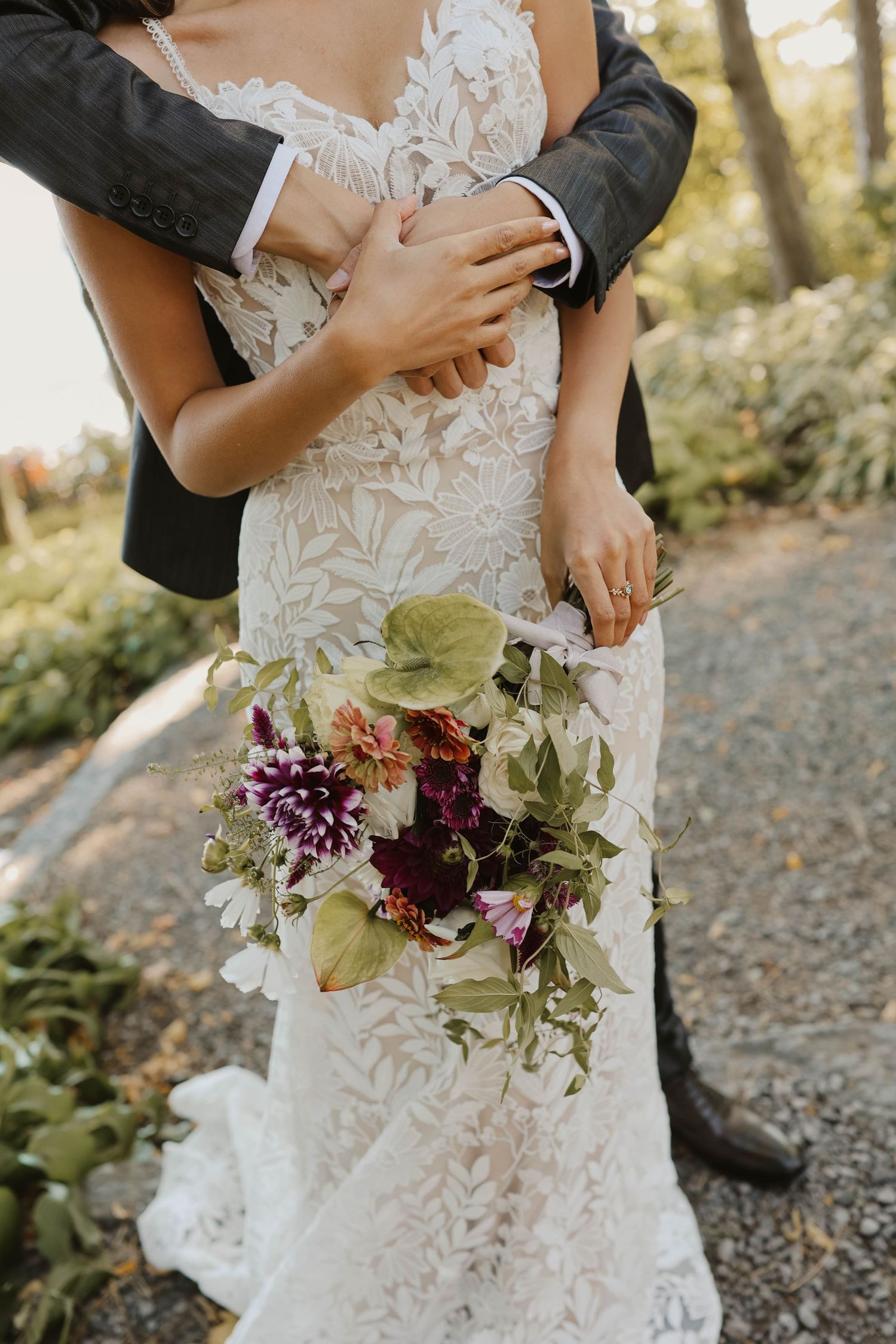 Groom embraces bride in a lace wedding gown while she holds a lush burgundy floral bouquet.