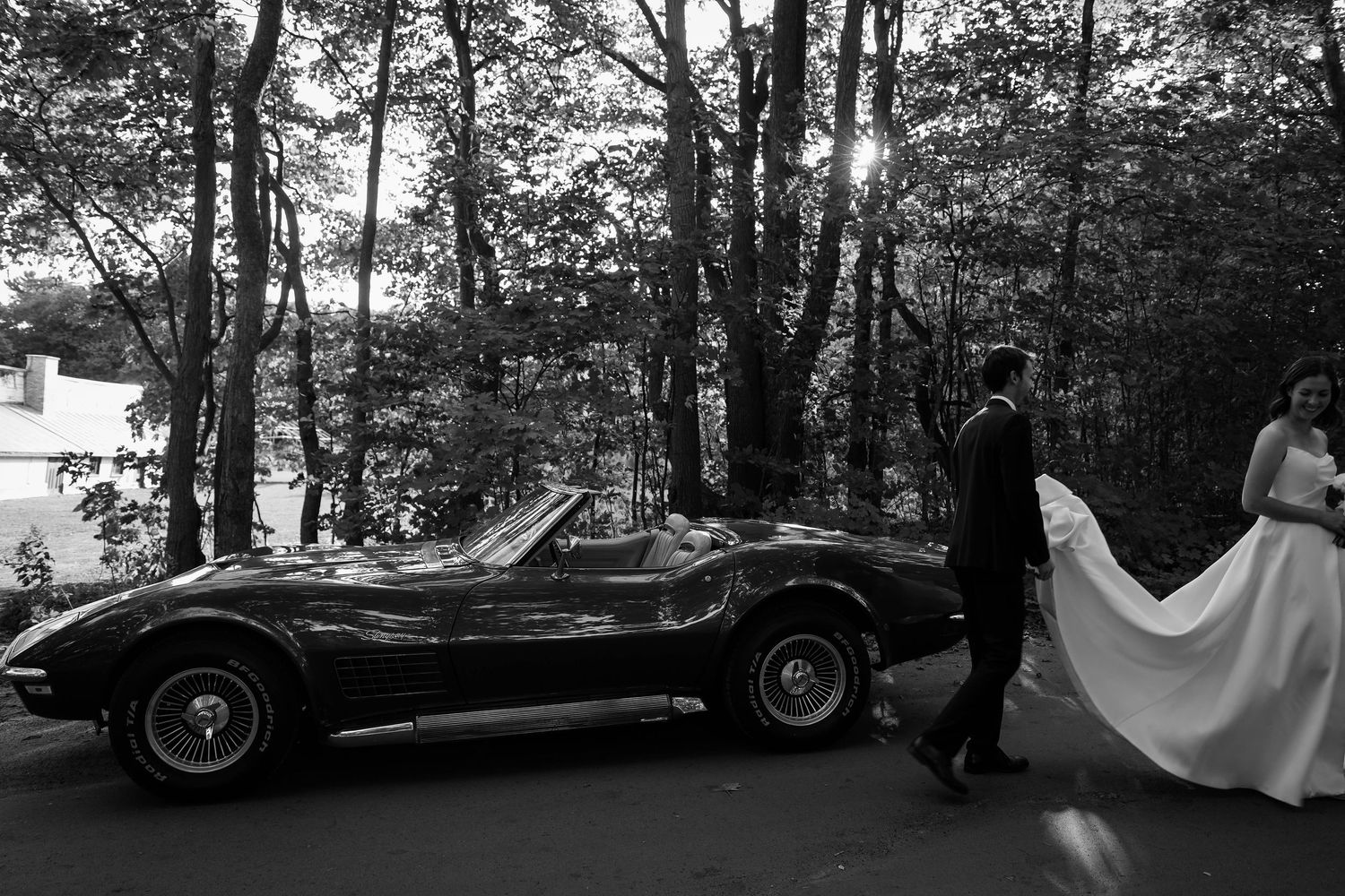 Black and white photo of bride and groom walking beside a vintage Corvette in a wooded setting.