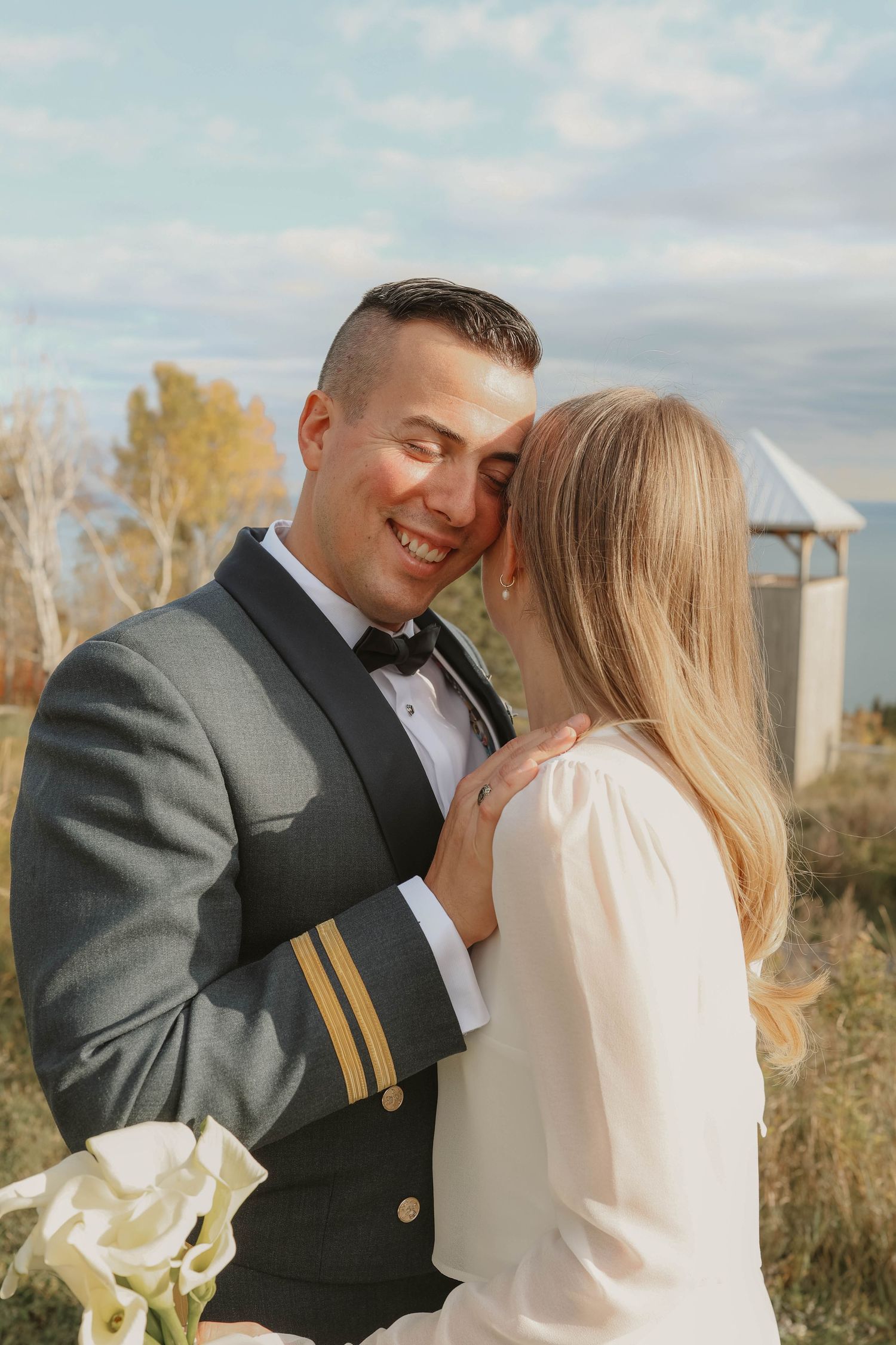 Smiling groom in military uniform embracing blonde bride outdoors on their wedding day.