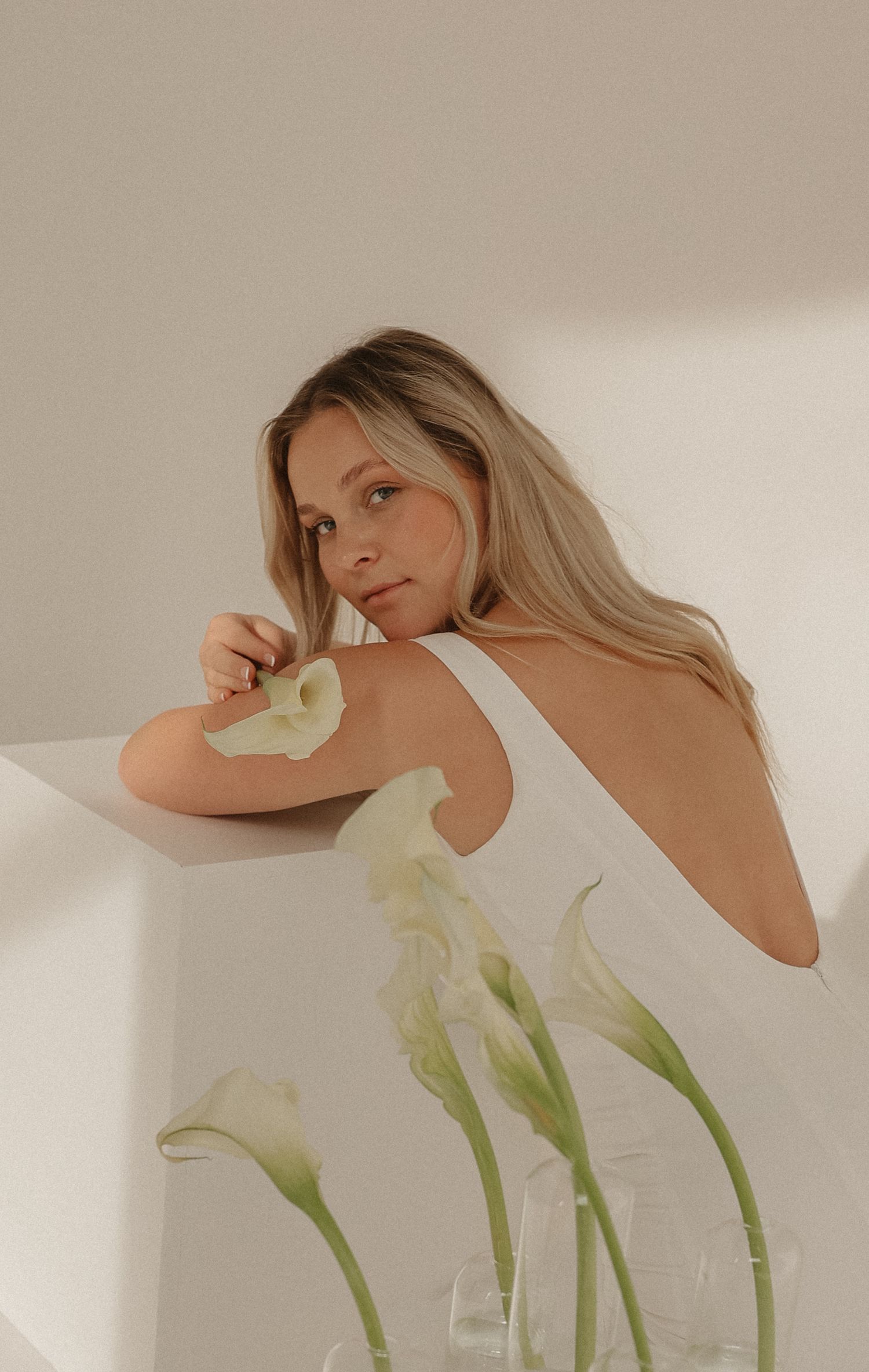 Blonde bride in backless white gown leaning on white surface with fresh calla lilies nearby.