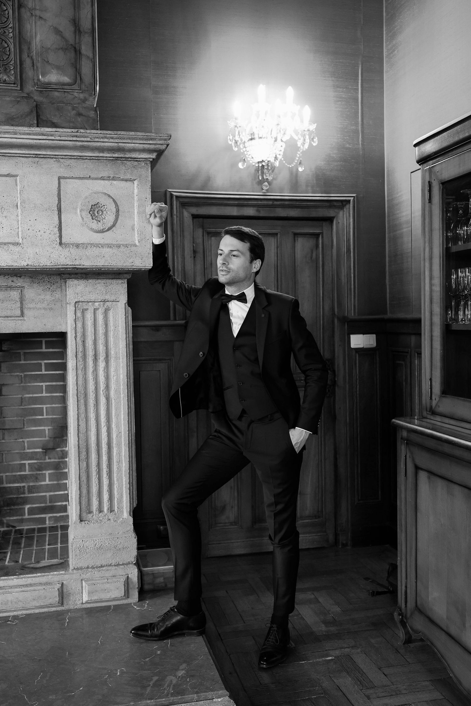 Sophisticated groom in black tuxedo posing by a fireplace in a classic interior with chandelier in black and white.