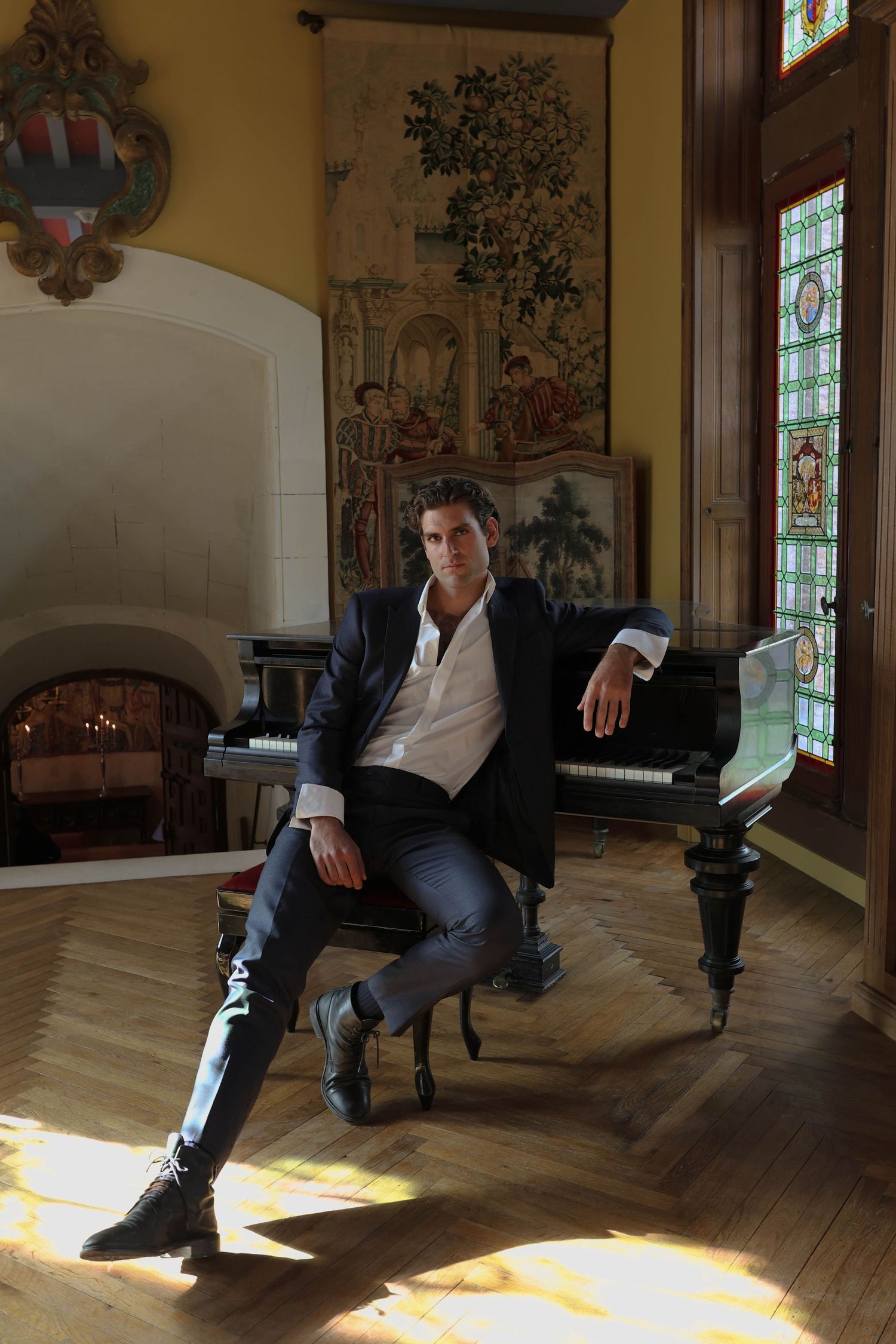 Confident man in dark suit seated at grand piano in ornate room with tapestry and stained glass windows.