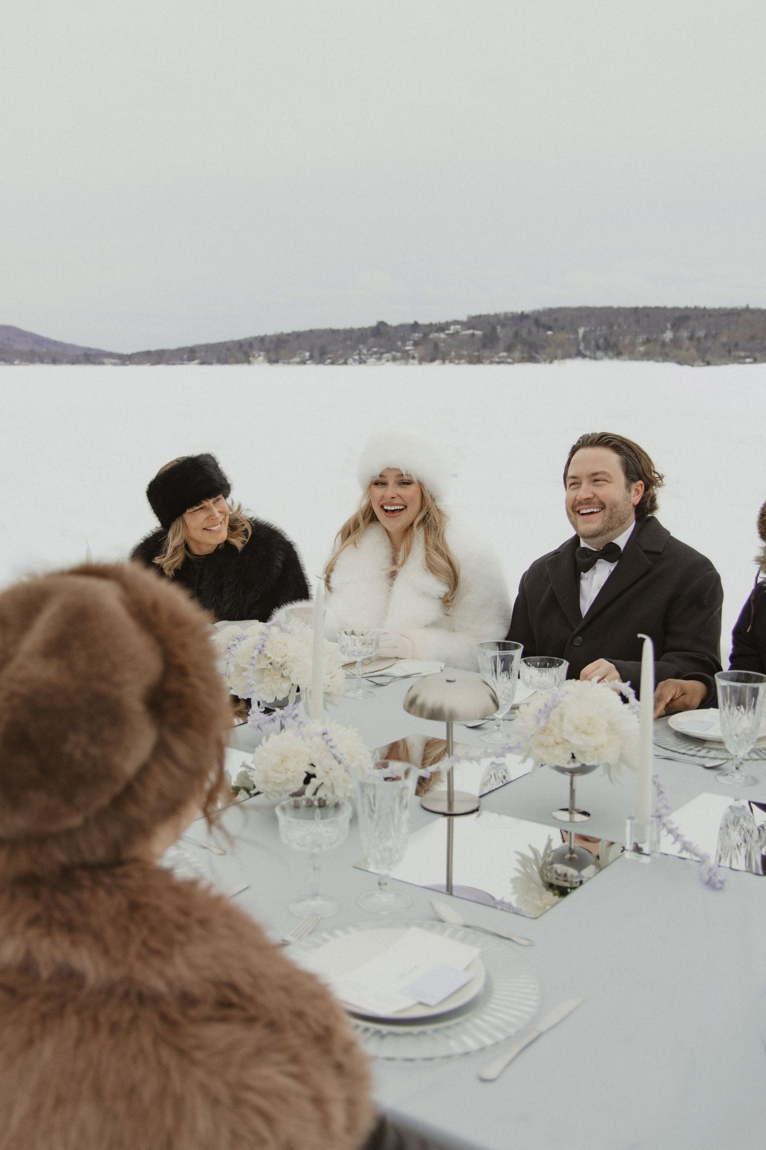 Bride and groom share joyful laughter at outdoor winter wedding reception on frozen lake.