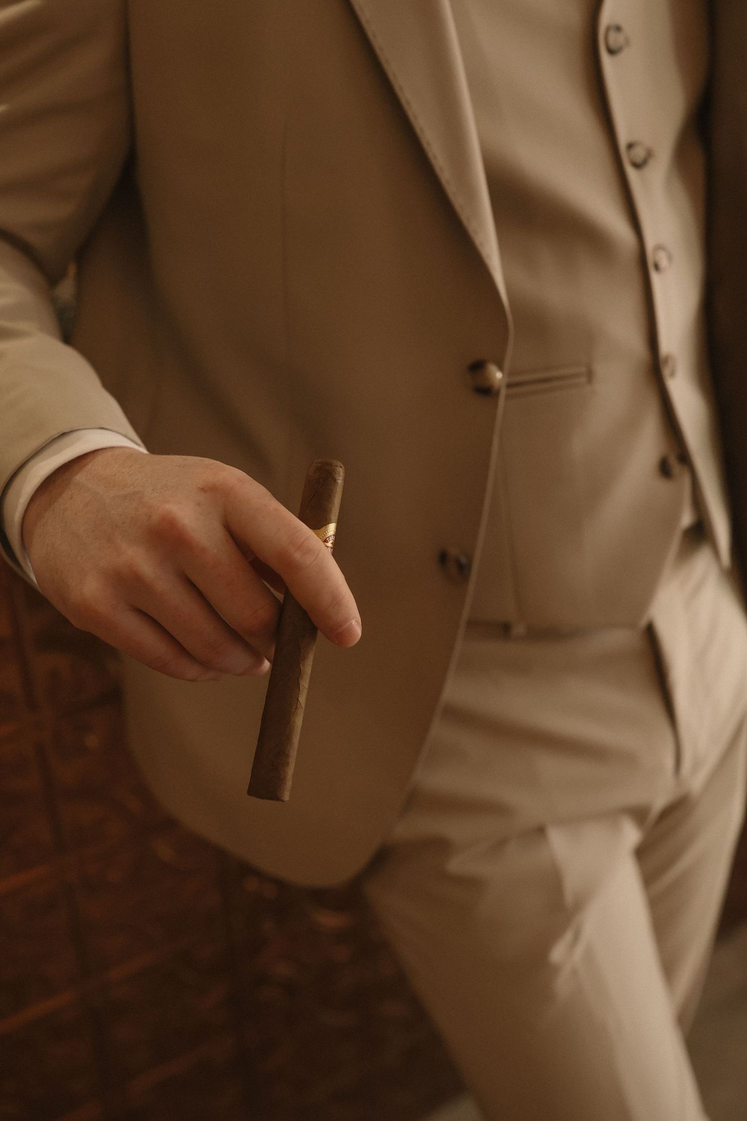 Man in beige three-piece suit holding a cigar, elegant formal attire with subtle earth tones.