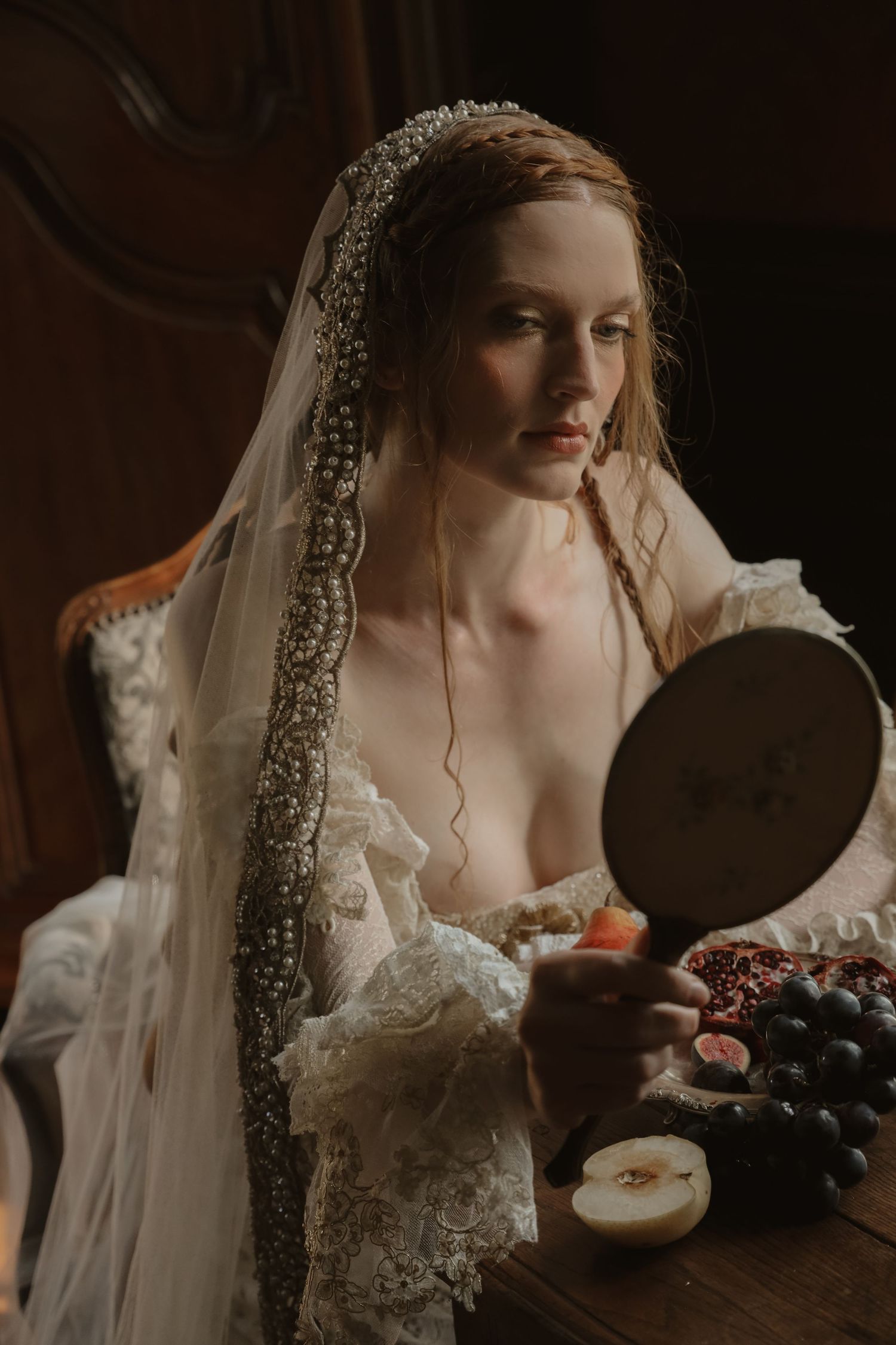 Bride in beaded veil holding mirror with pomegranate and grapes in Renaissance-inspired portrait.