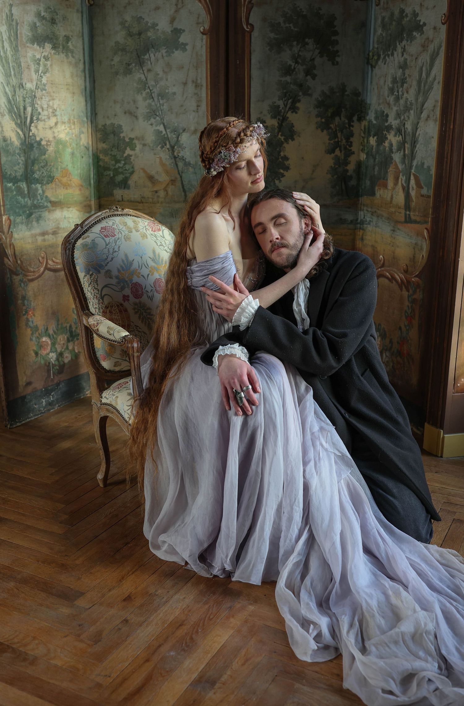 Couple in Renaissance-inspired attire pose intimately in ornate room with tapestry wall panels.