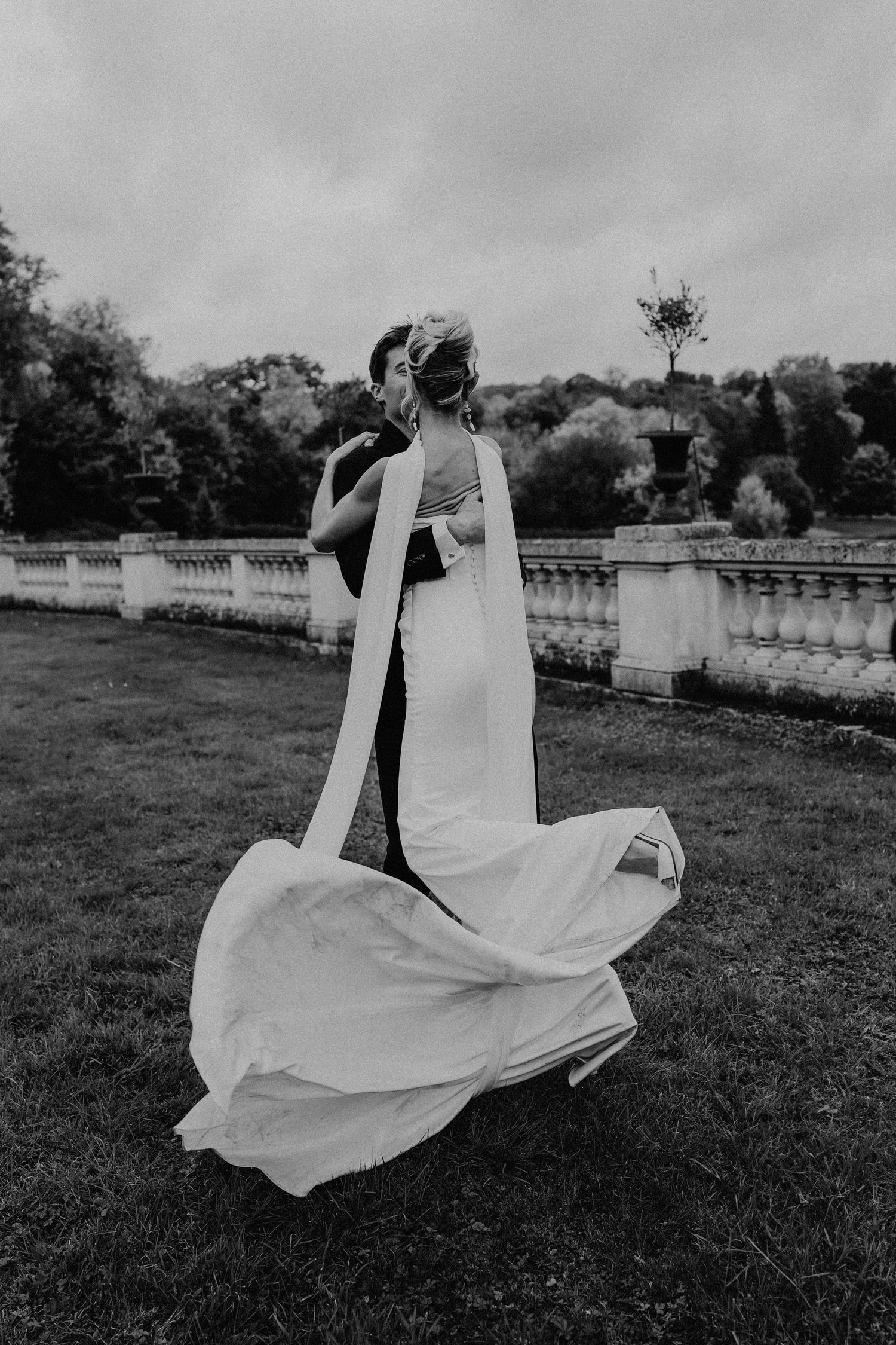 Black and white photo of bride and groom dancing on lawn, wedding dress flowing in the wind.