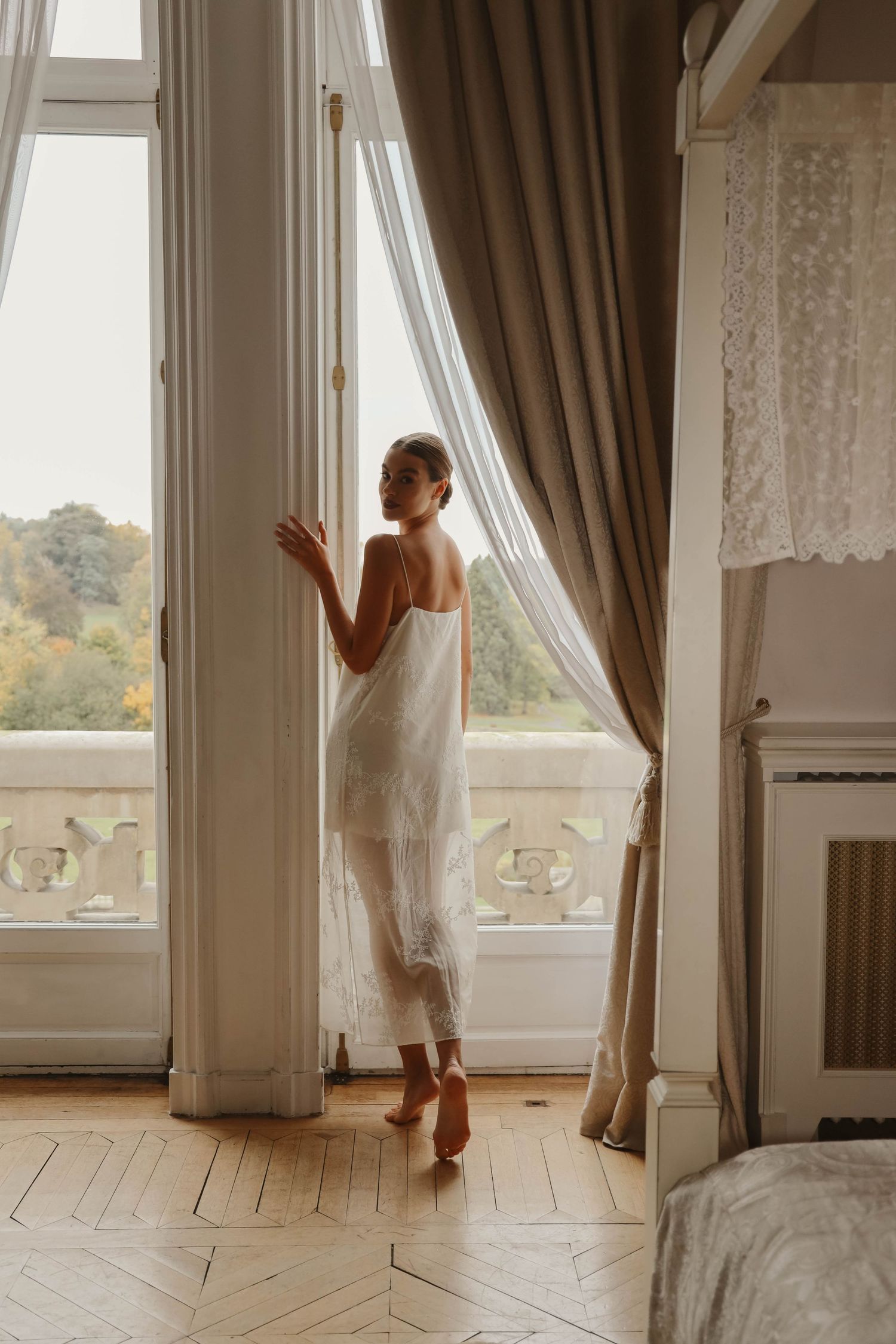 Elegant bride in a sheer white slip dress standing by tall French windows with flowing curtains in a chateau.