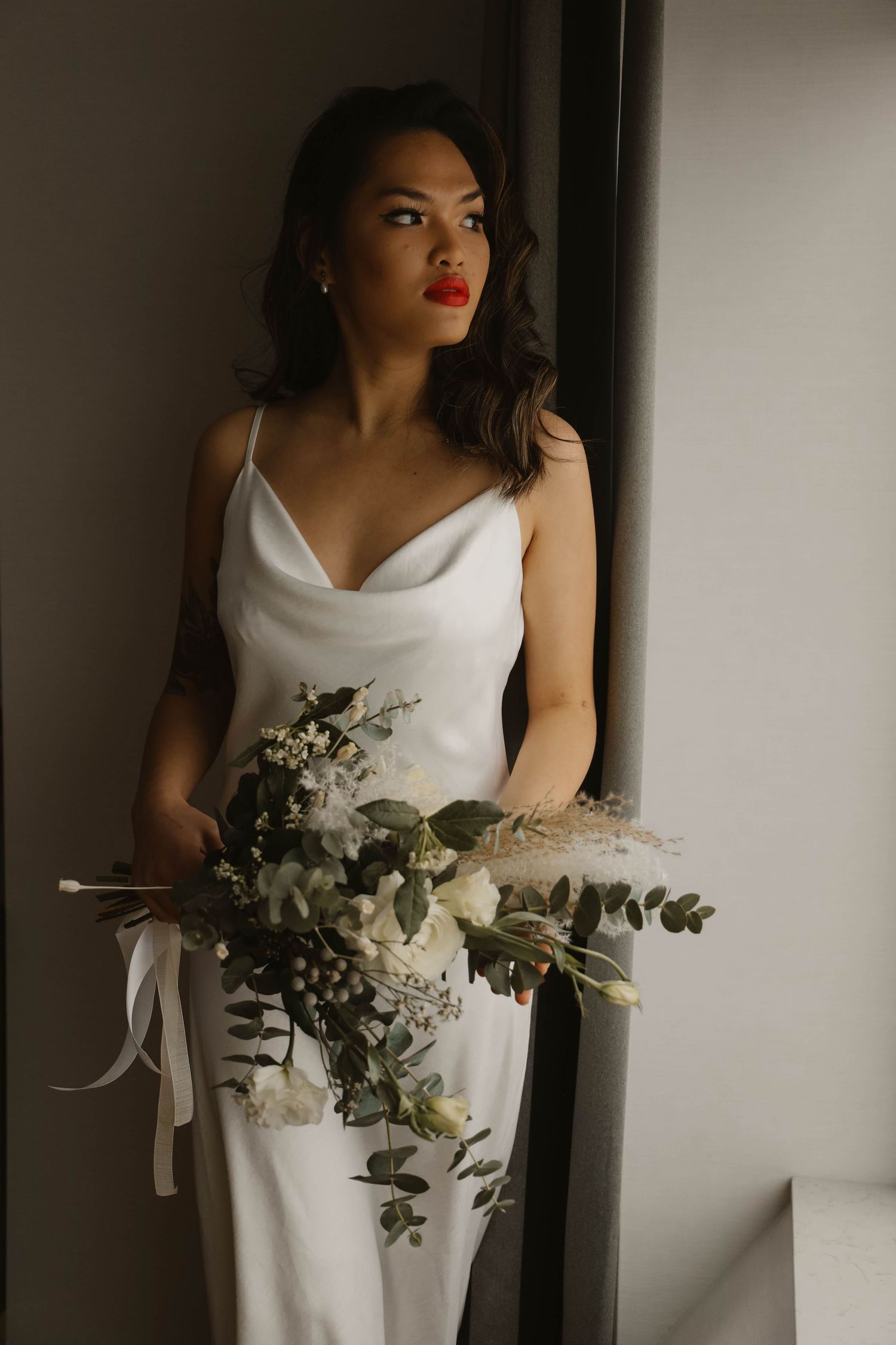 Elegant bride in white satin slip dress holding a lush bouquet of white flowers and greenery.