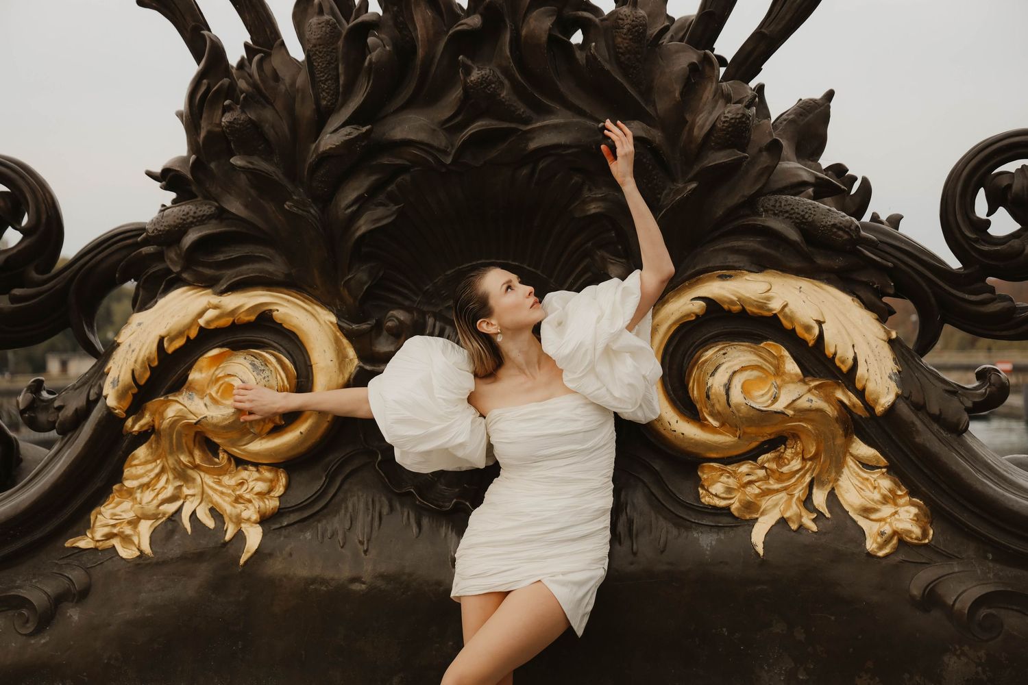 Woman in short white puff-sleeve bridal dress posing dramatically against ornate gold sculpture.