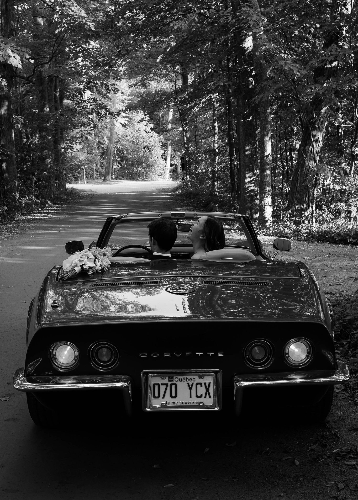 Black and white photo of a couple in a classic Corvette convertible on a tree-lined road.