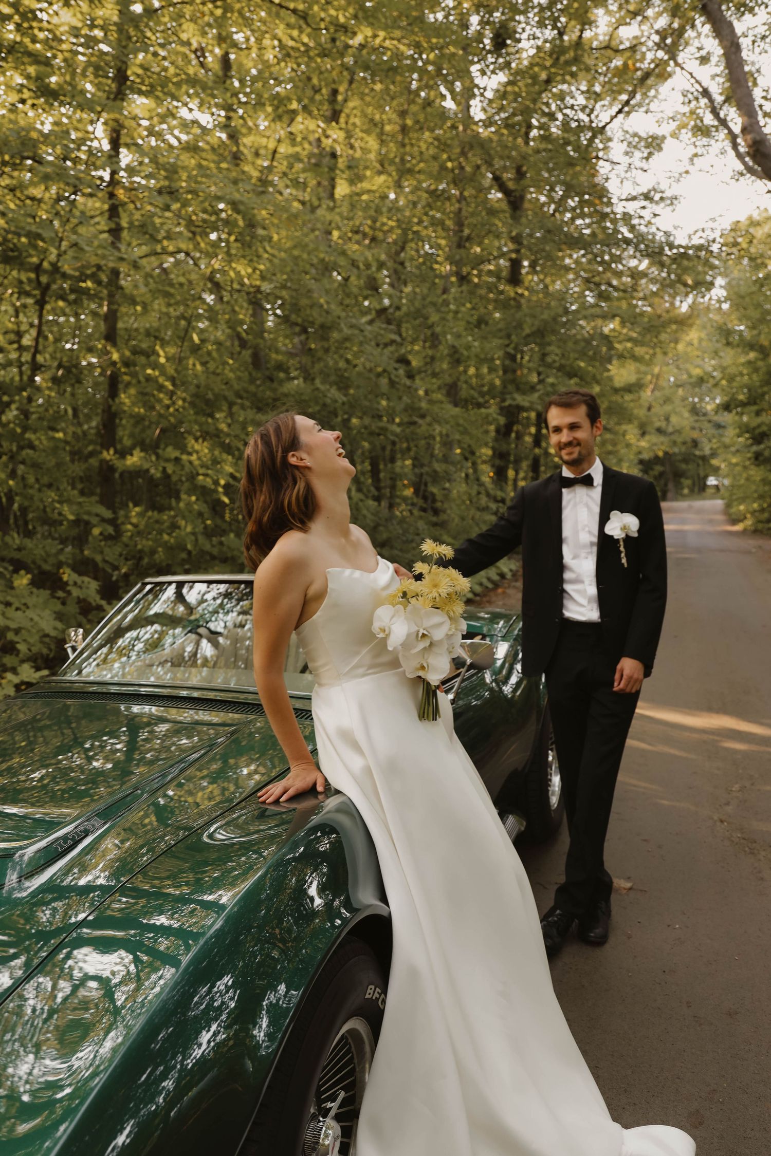 Laughing bride in white gown with yellow bouquet leaning on vintage green Corvette with groom.