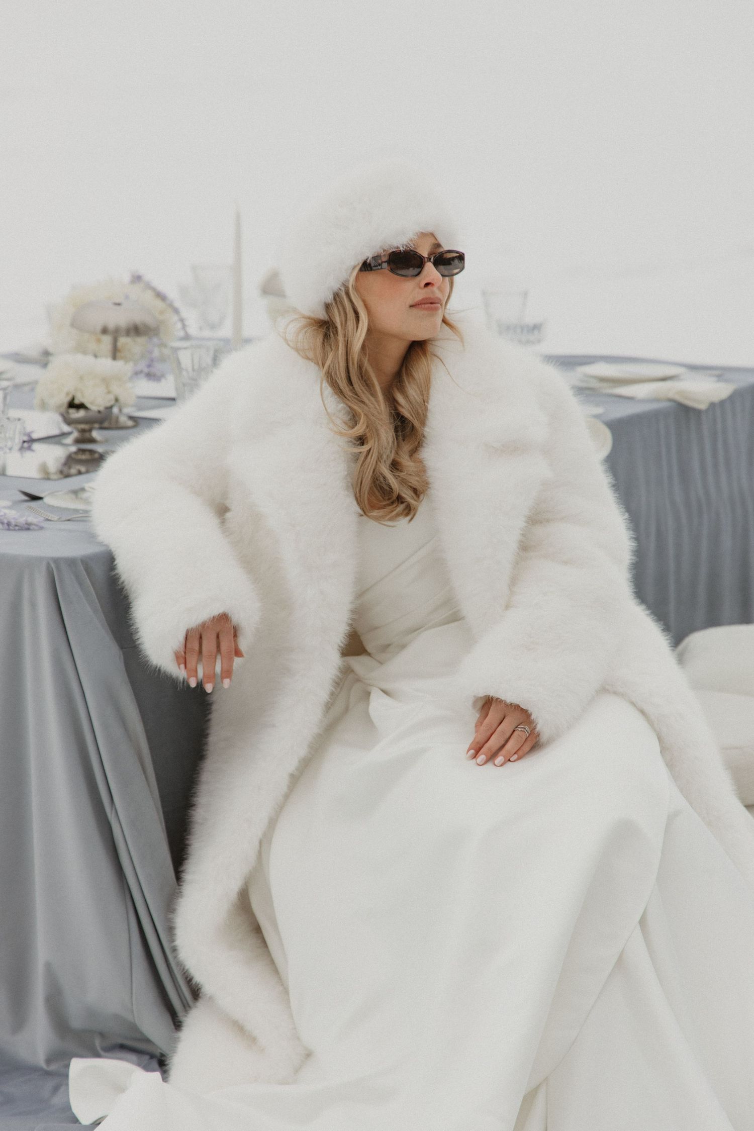 Elegant bride in white faux fur coat and hat poses at winter wedding reception table.