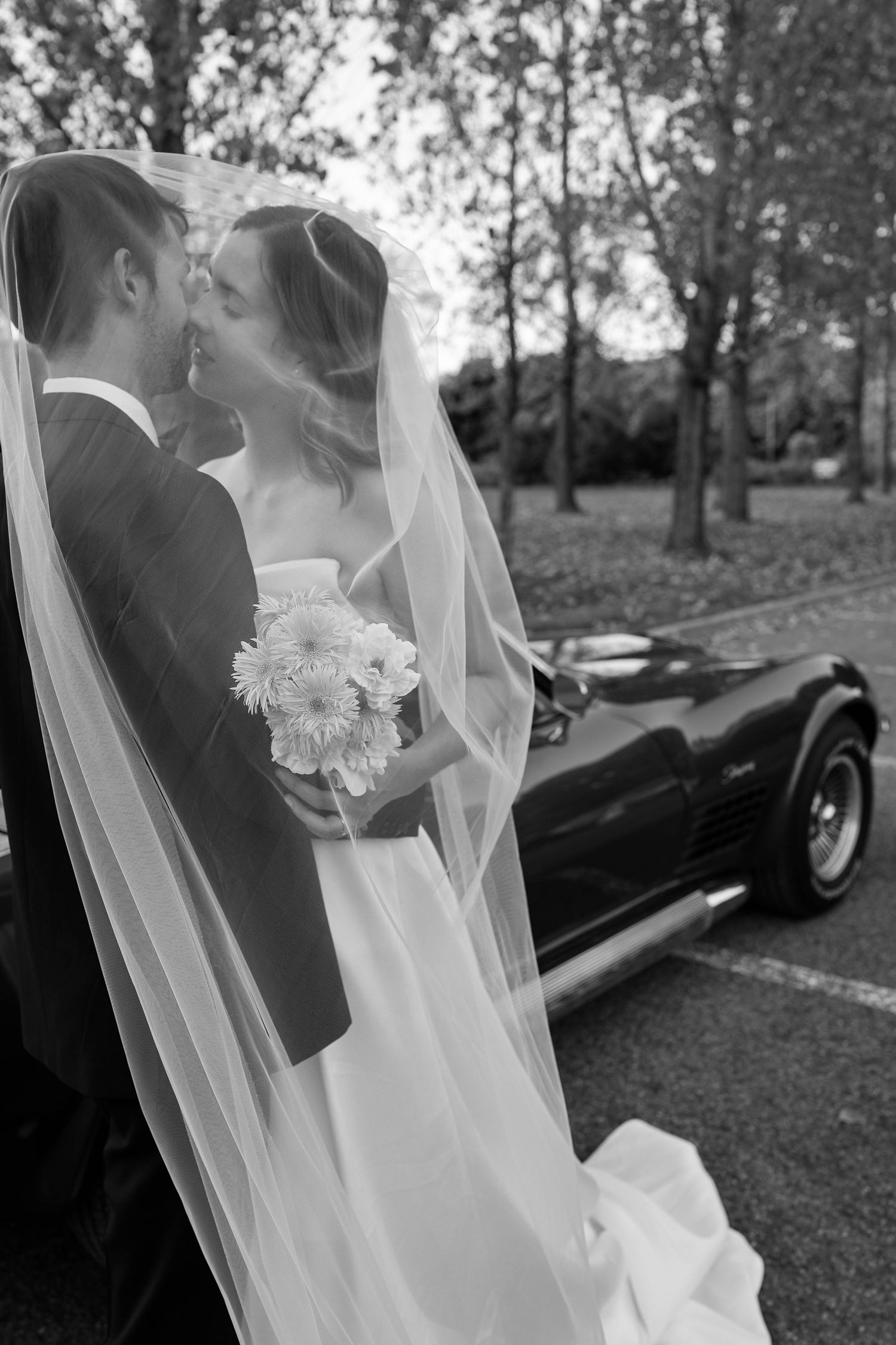 Bride and groom sharing a kiss under wedding veil beside vintage black convertible car in black and white.