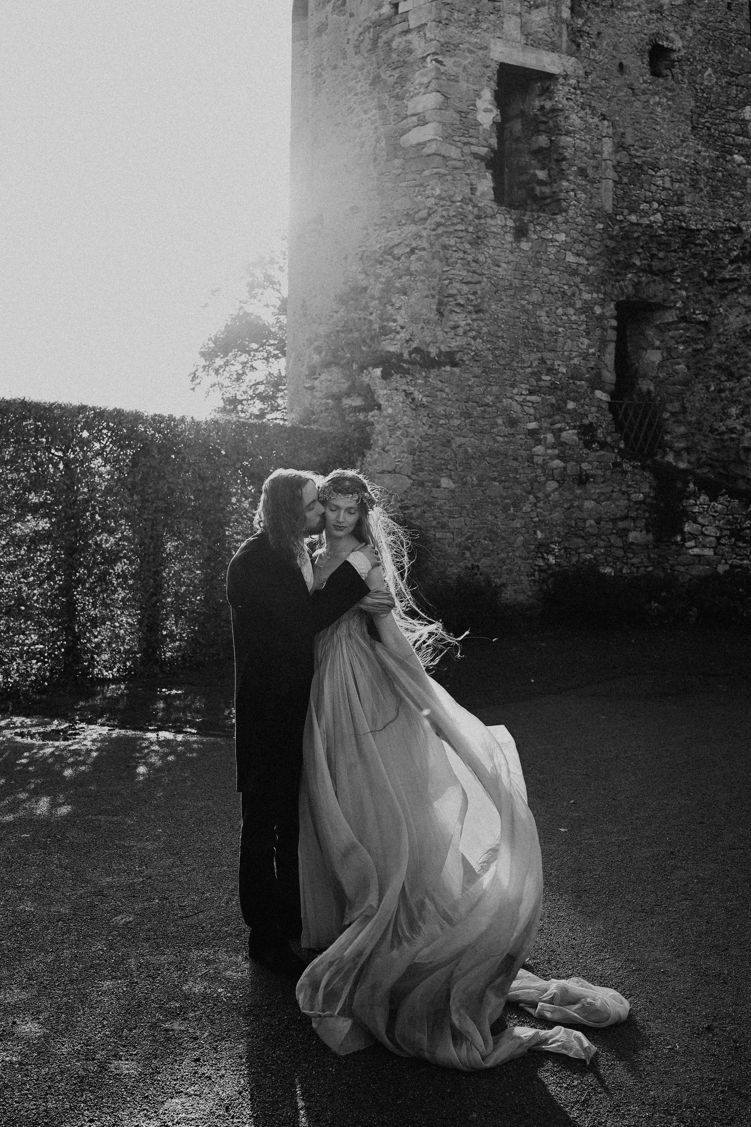 Black and white photo of couple embracing in flowing wedding attire outside a stone castle ruin.