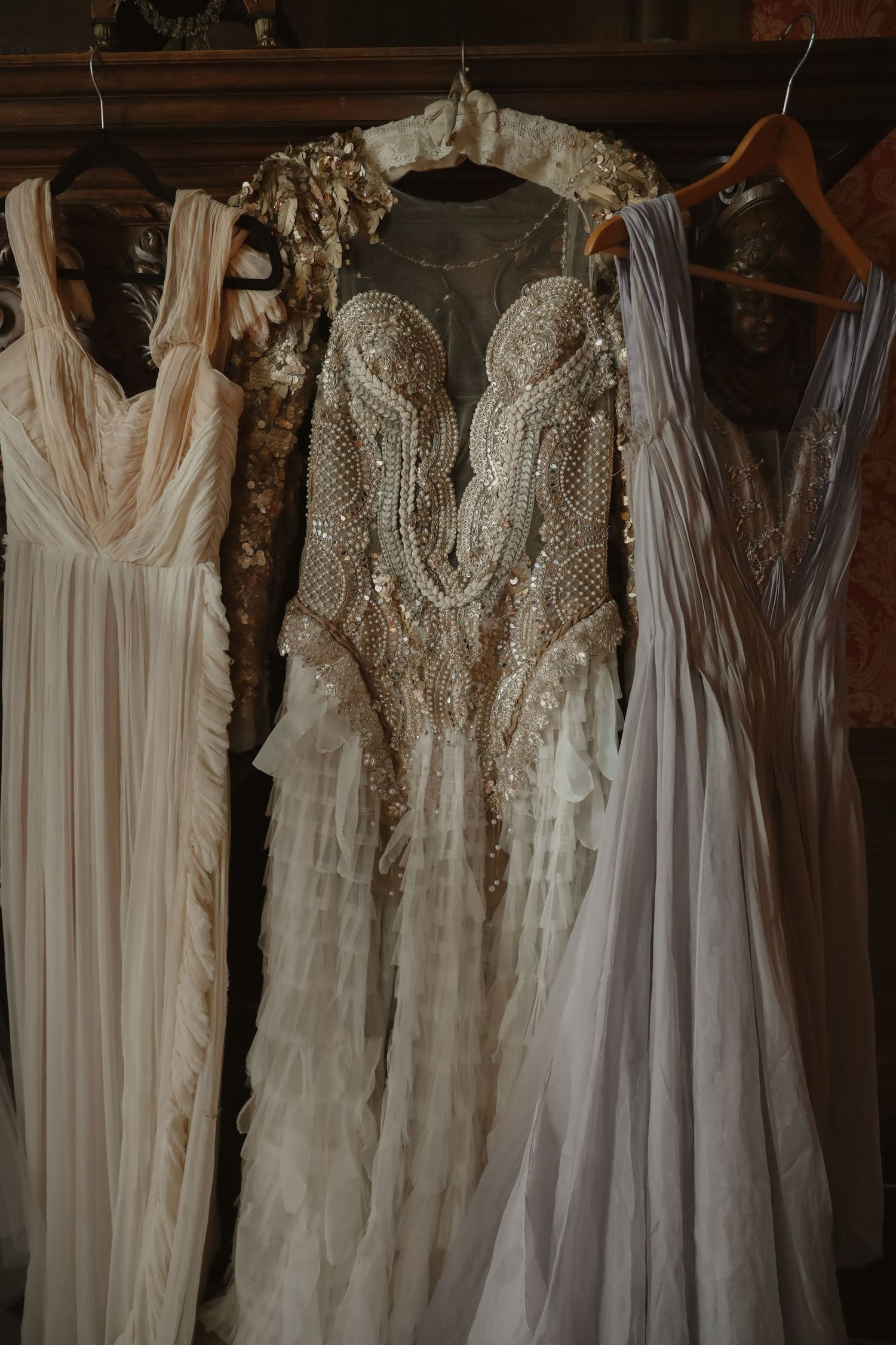 Ornate beaded wedding dress with pearl embroidery displayed on hanger alongside white gowns.