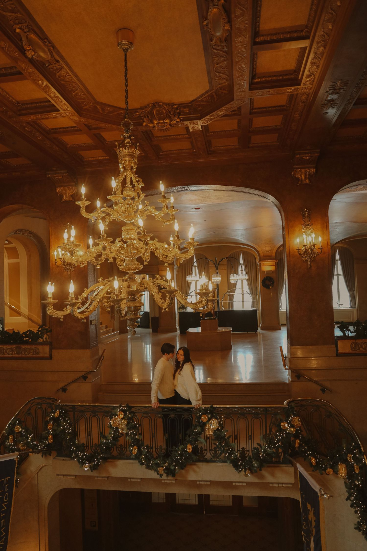 Couple embracing beneath a grand golden chandelier in an ornate historic ballroom.