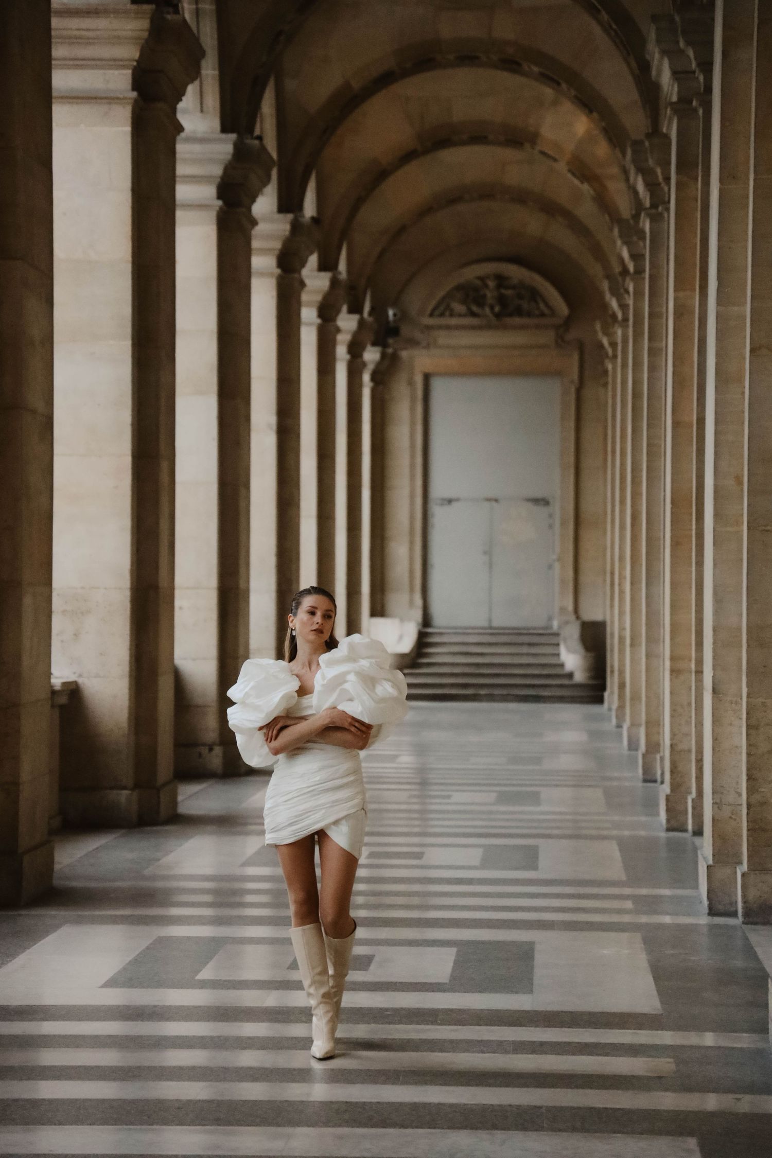 Woman in white ruffled mini dress and boots poses in elegant Parisian arcade with arched columns.