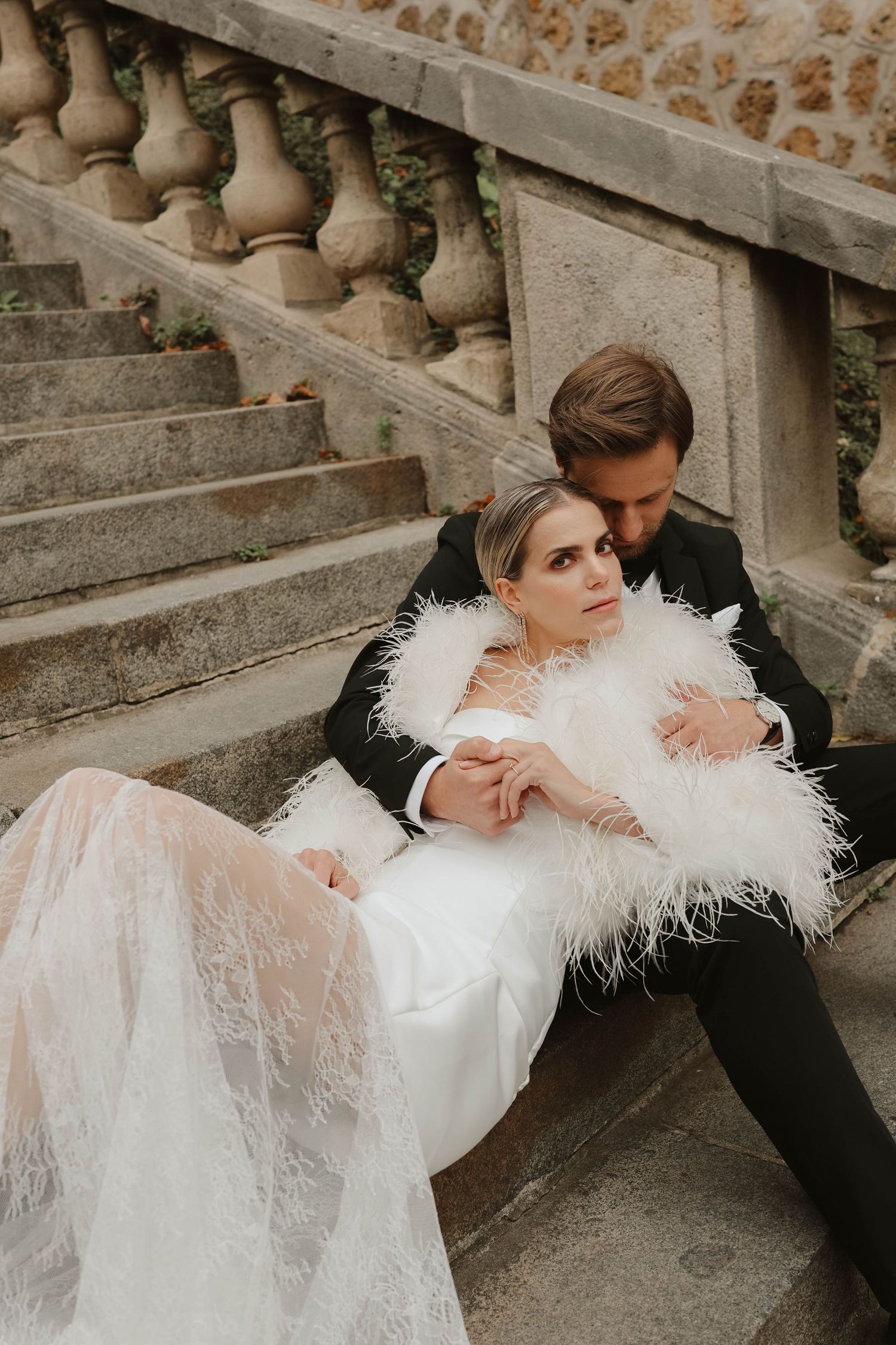 Elegant bride and groom sitting on stone steps, bride wearing a white feather wrap.