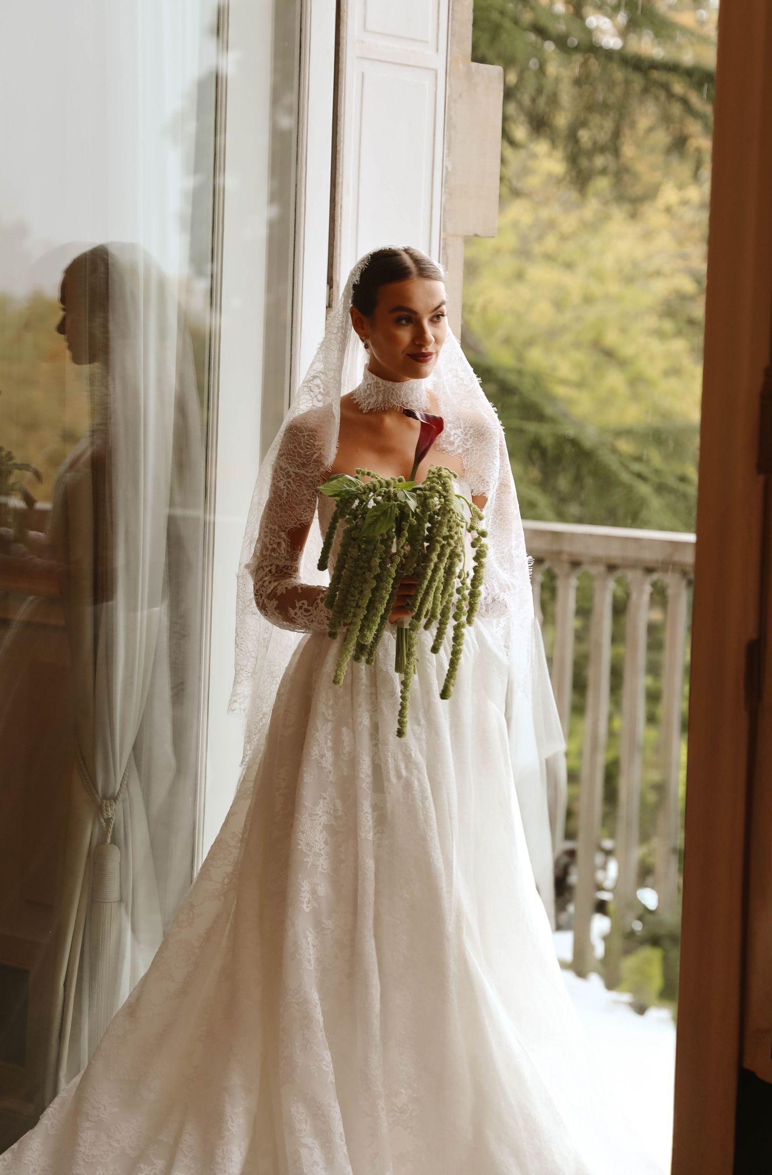 Bride in strapless lace wedding dress with veil holding cascading green bouquet by glass doors.