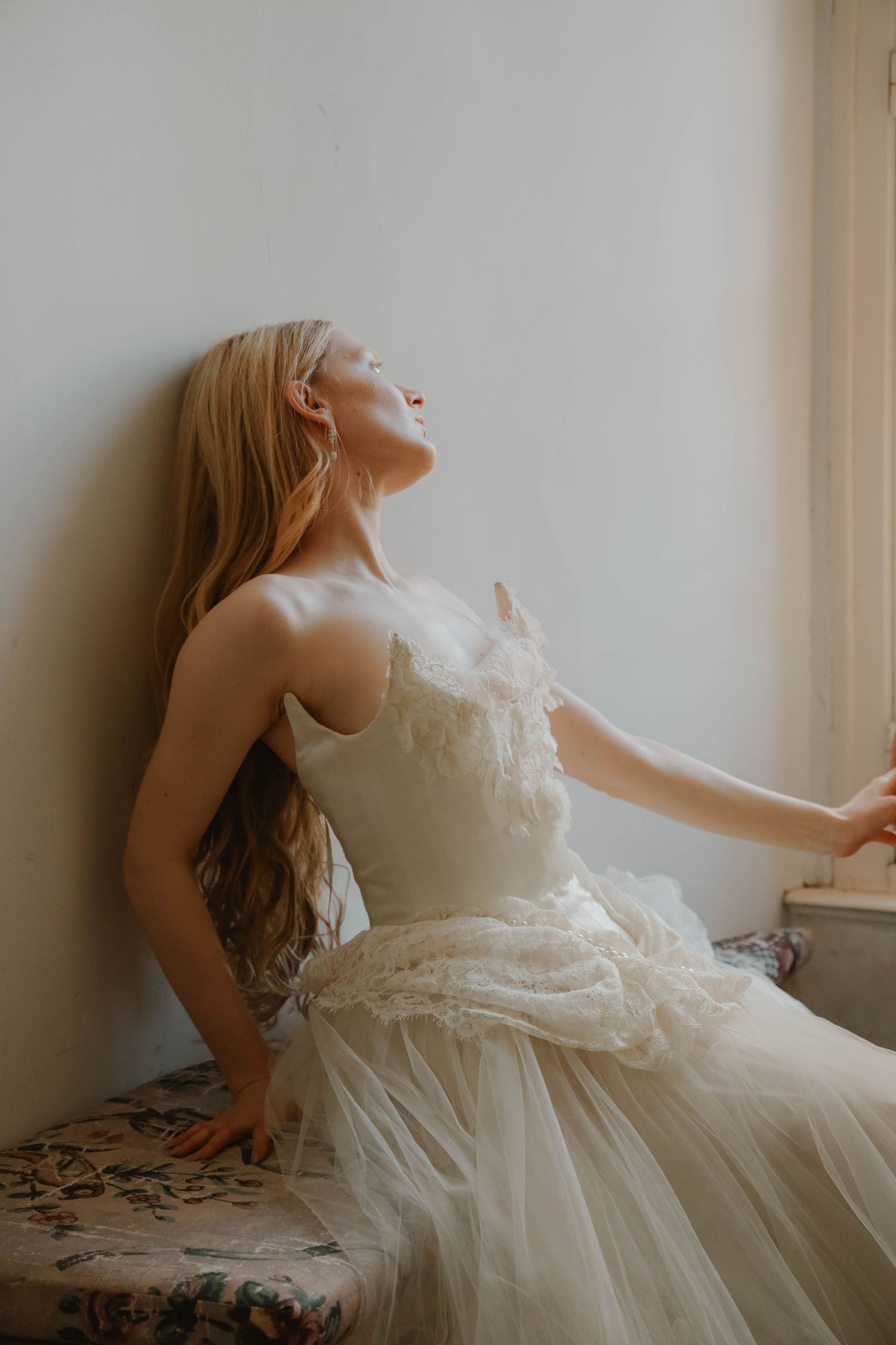Dreamy bride in a delicate tulle wedding gown sitting by a window with flowing blonde hair and eyes closed.