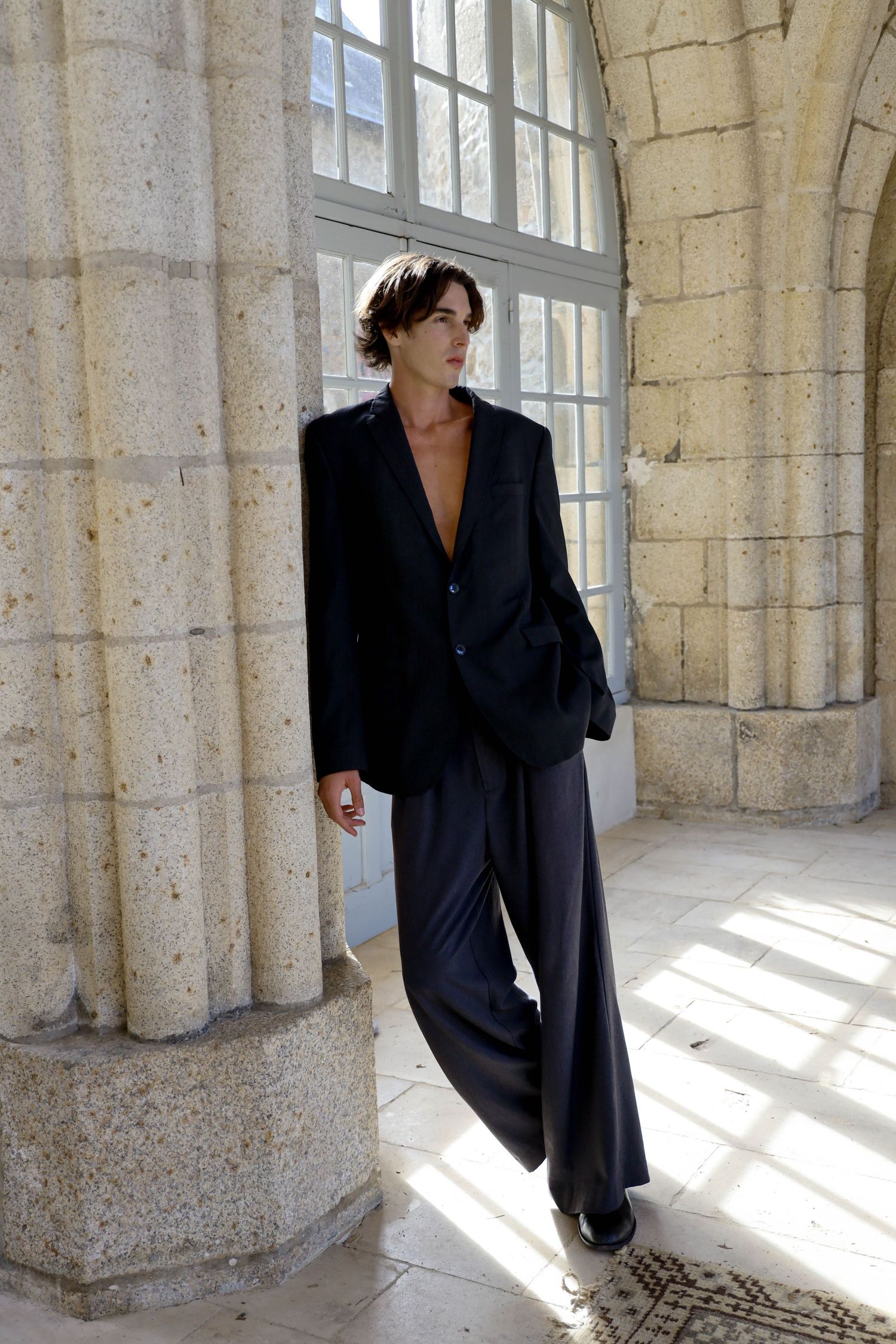 Stylish person in dark oversized blazer and wide-leg trousers leaning against stone column.