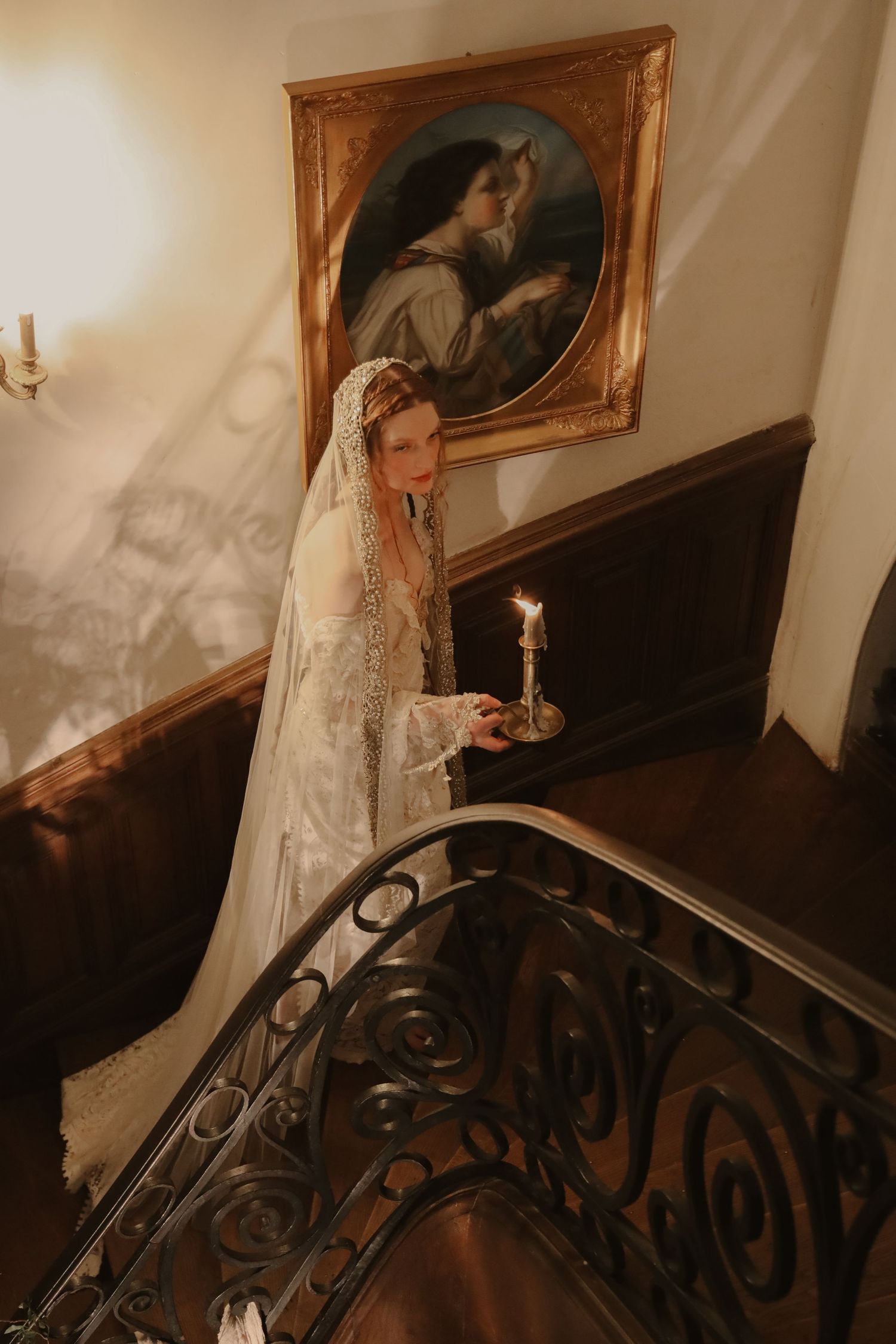 Bride in lace gown and veil holding candle on ornate staircase beside classic portrait.