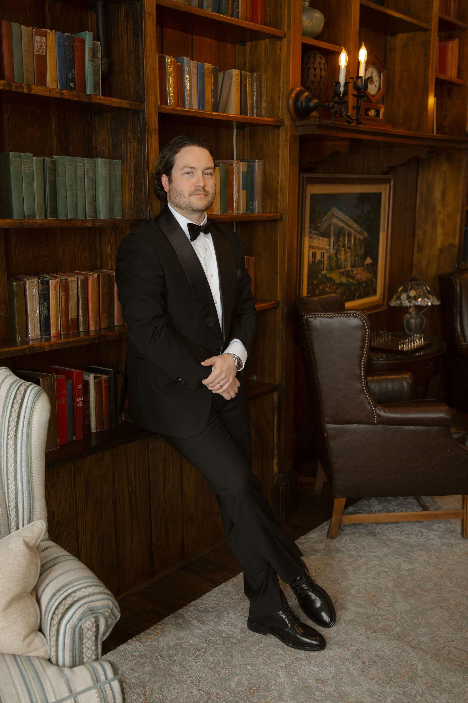 Sophisticated groom in black tuxedo with bow tie posing in elegant wood-paneled library.