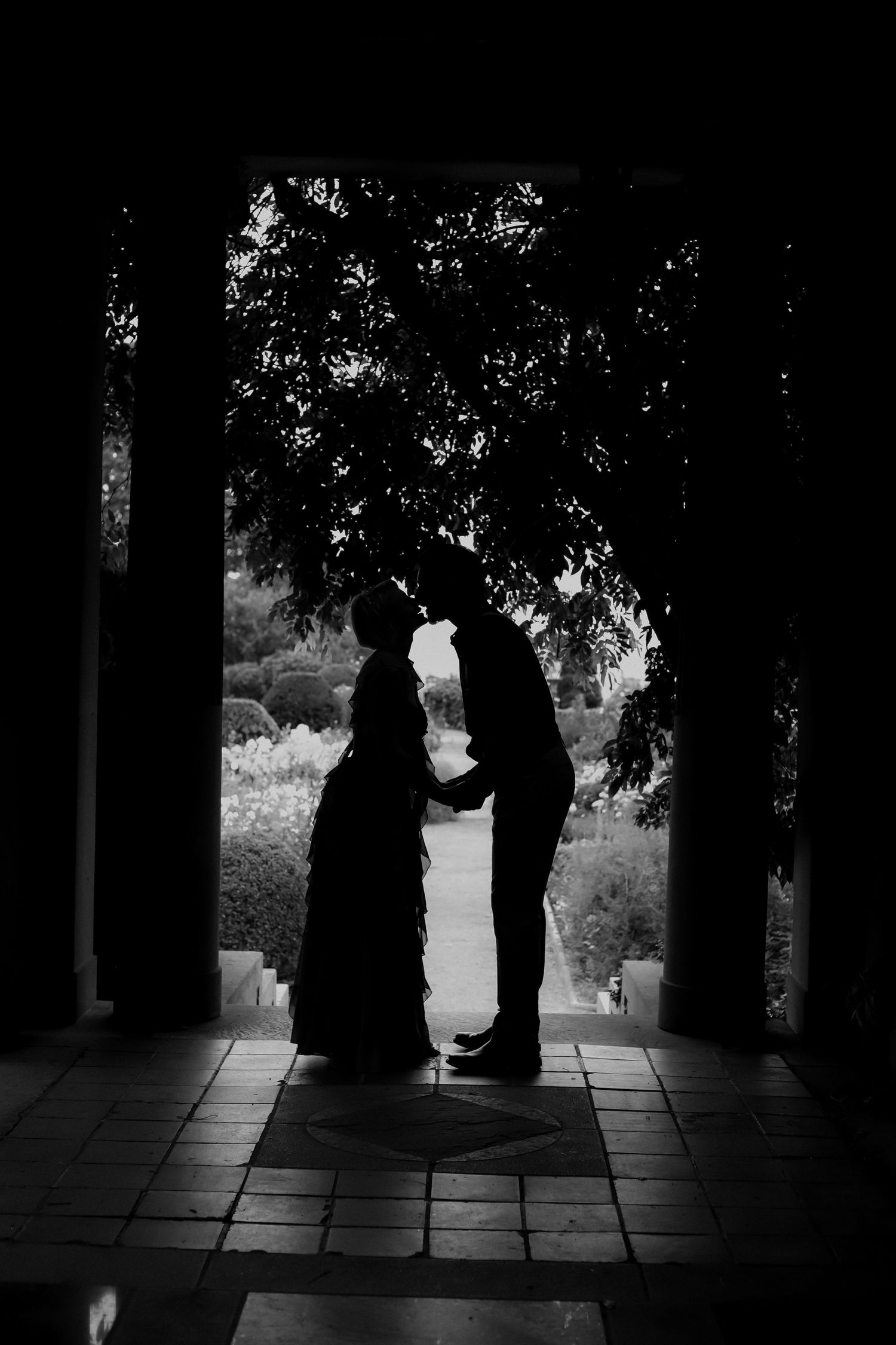 Black and white silhouette of a couple holding hands in a garden archway, framed by lush trees.