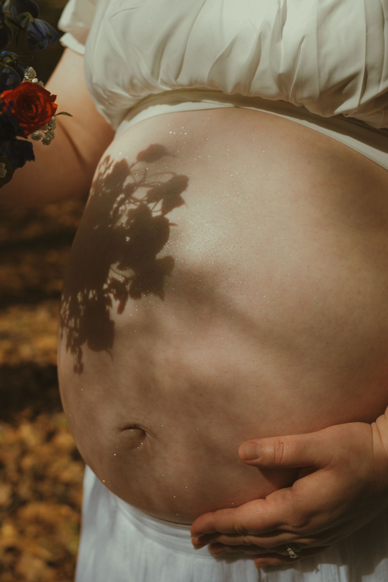 Close-up of pregnant woman's bare belly with floral shadow, wearing white dress and holding colorful bouquet outdoors.