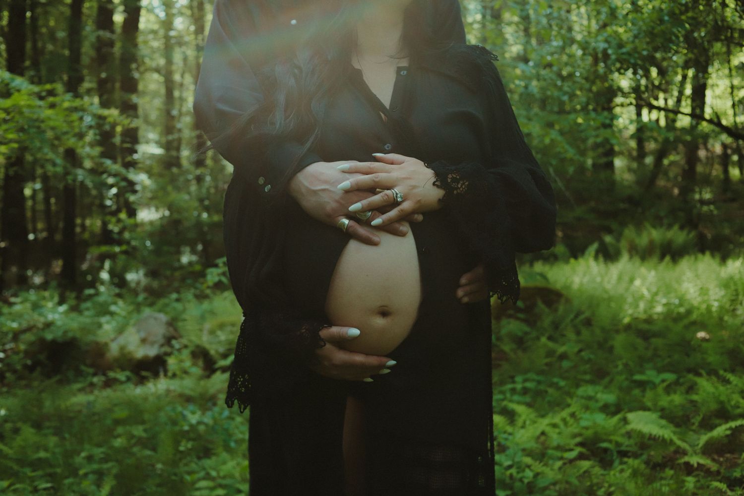 Pregnant woman in black dress cradling baby bump in lush green forest setting.