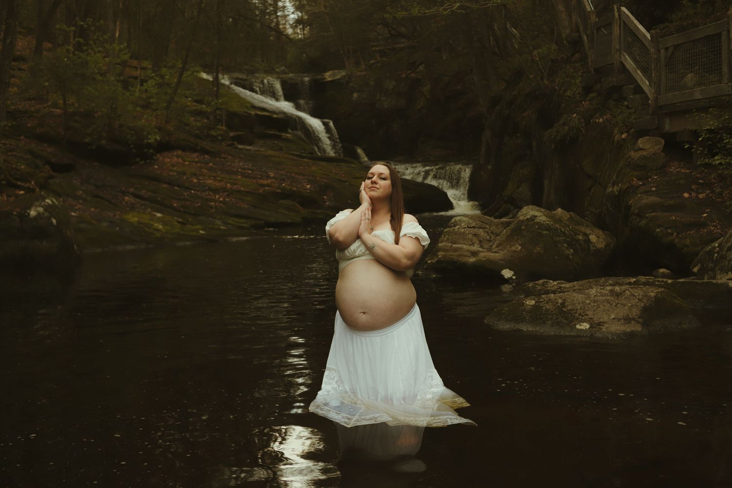 Pregnant woman in white dress standing in a forest stream near a waterfall during a maternity photo shoot.