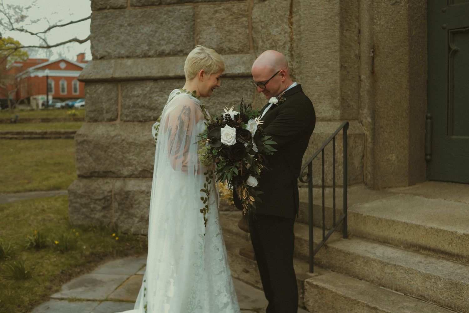 Bride in a white lace cape veil and groom in a black tuxedo bowing heads in prayer outside a stone church.
