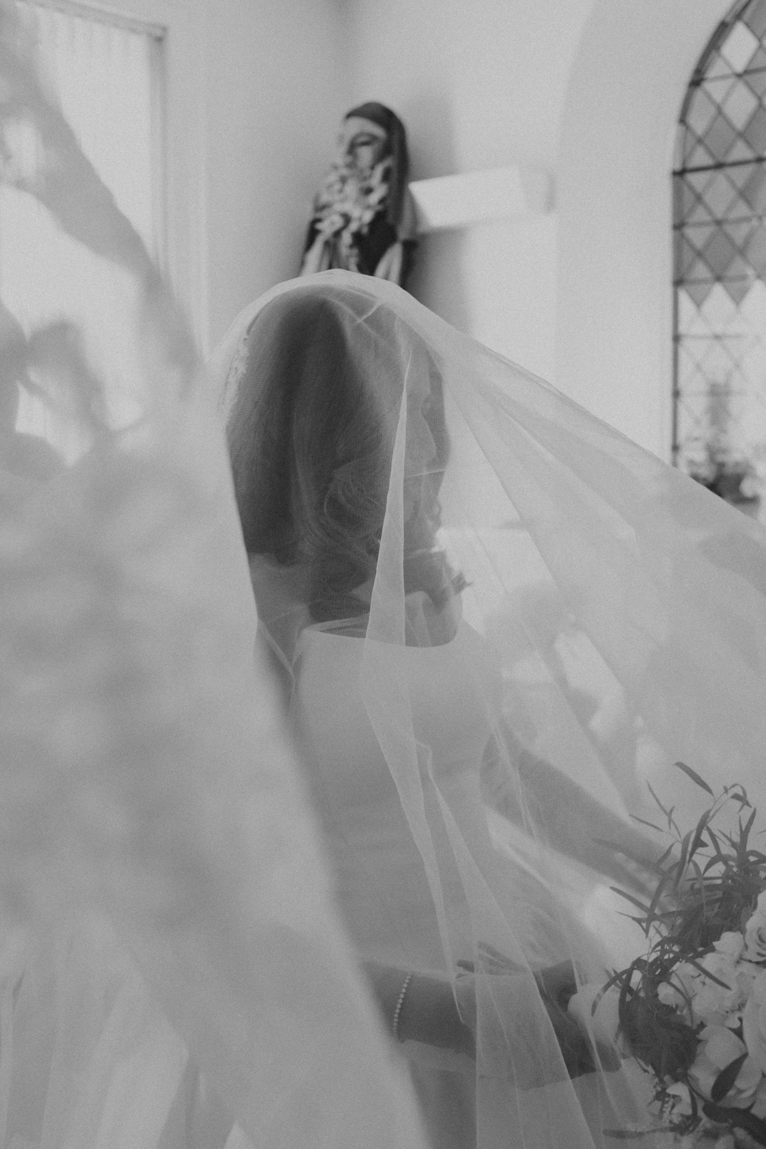 Black and white bridal portrait with flowing veil draped elegantly in a softly lit church setting.