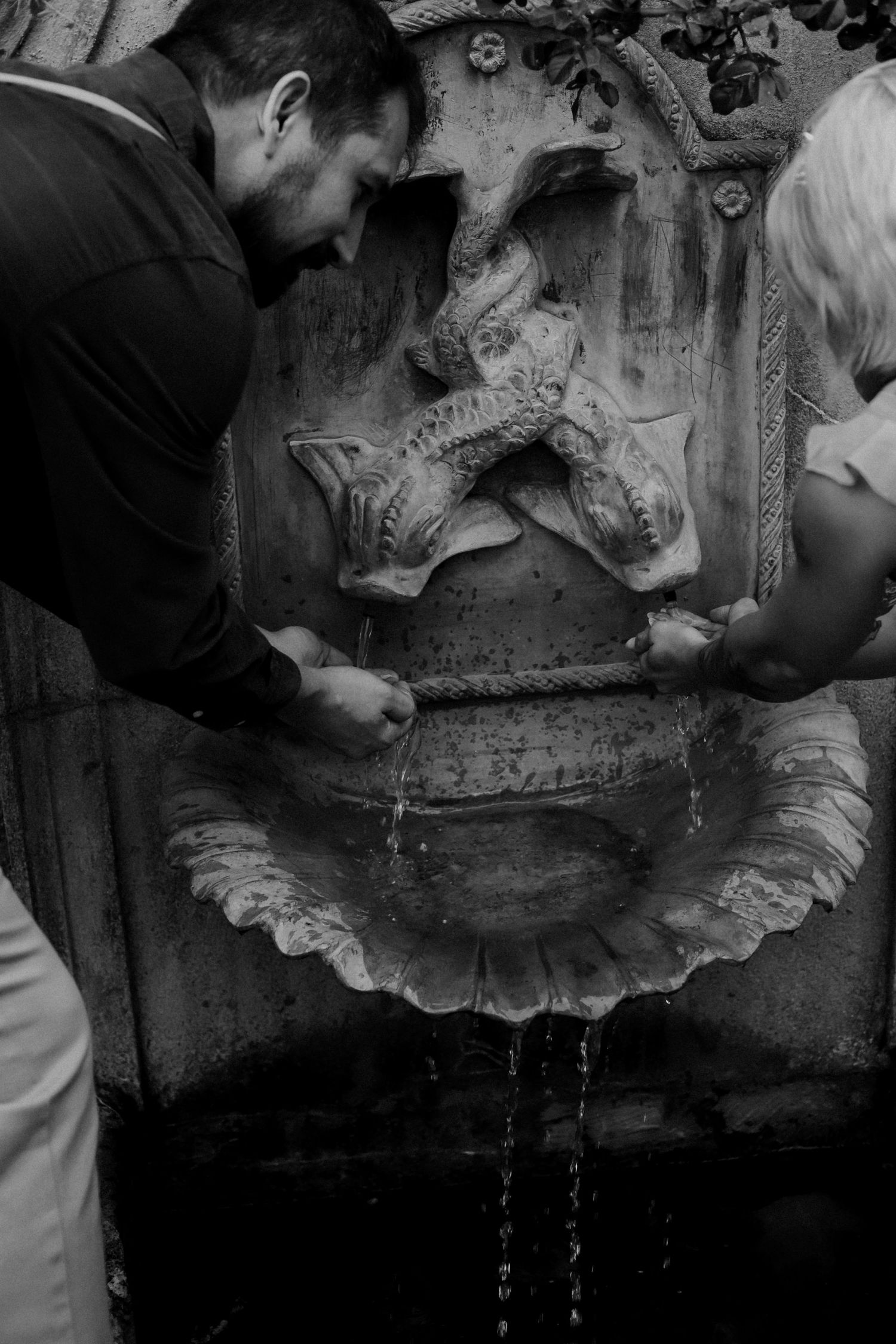 Two people drinking from an ornate stone fountain with fish sculptures in black and white photography.