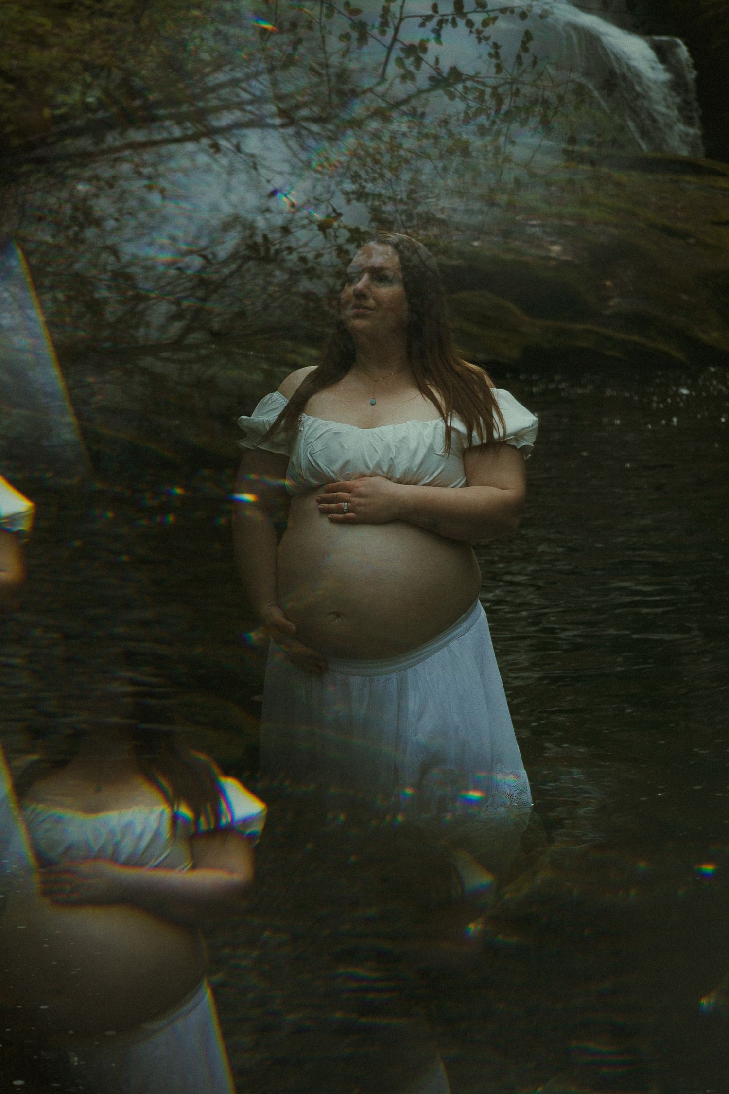 Pregnant woman in white dress standing in a forest stream with dreamy light effects and waterfall backdrop.